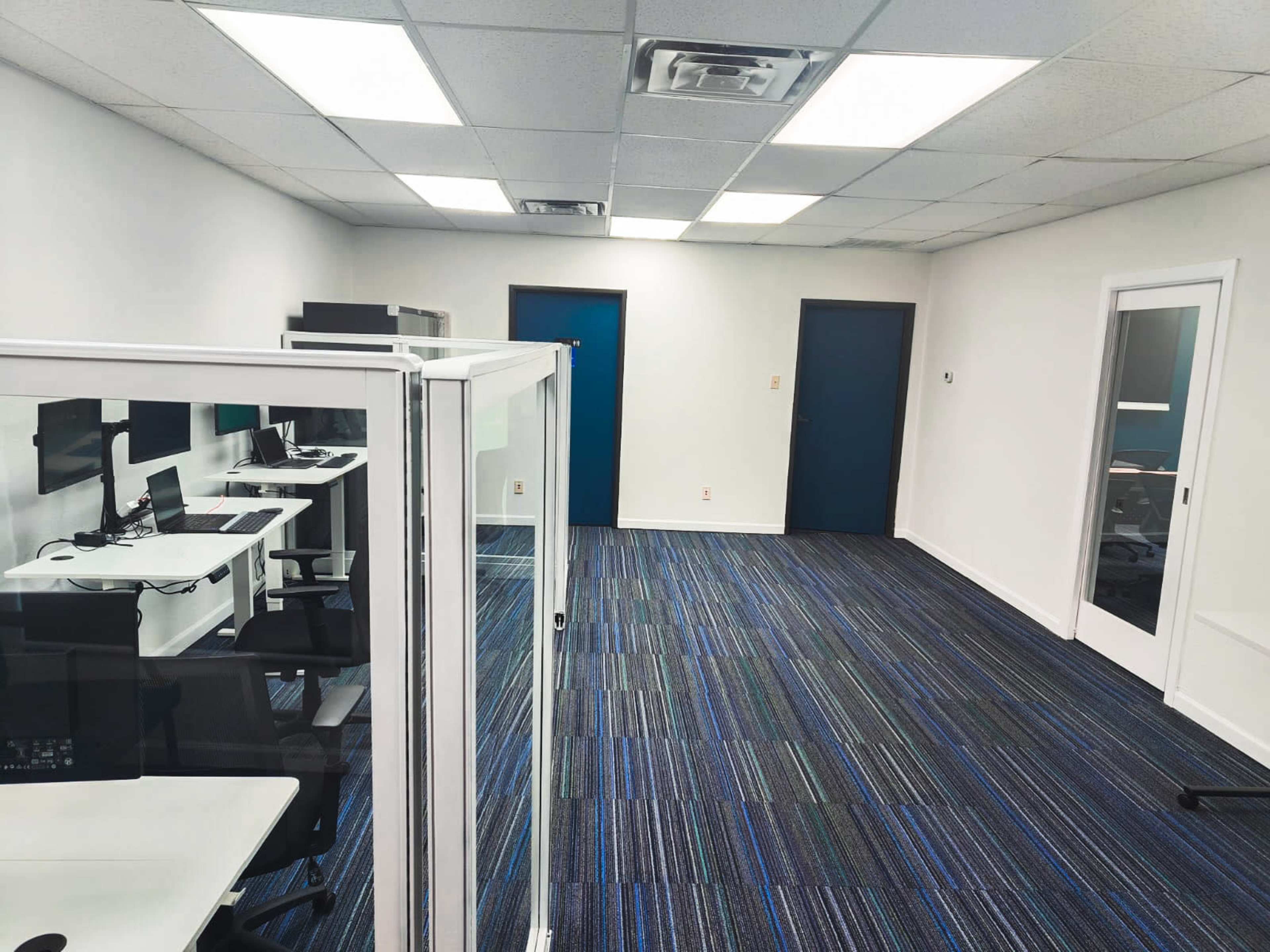 A modern office space with cubicles, desks, and blue carpet, featuring white walls and dark blue doors.