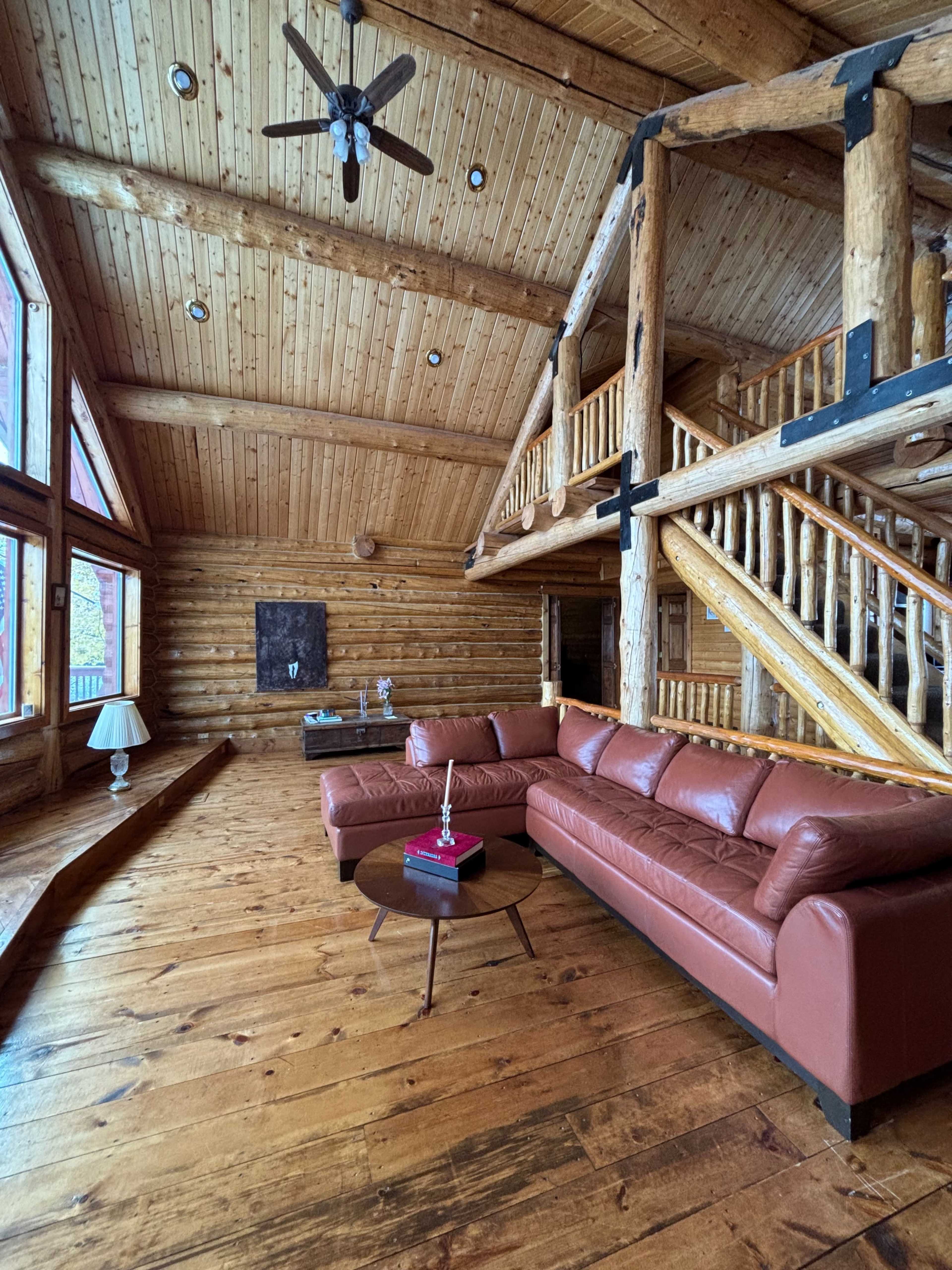 The image shows a spacious log cabin interior featuring a large leather sofa, wooden beams, and expansive windows that allow natural light to fill the room.