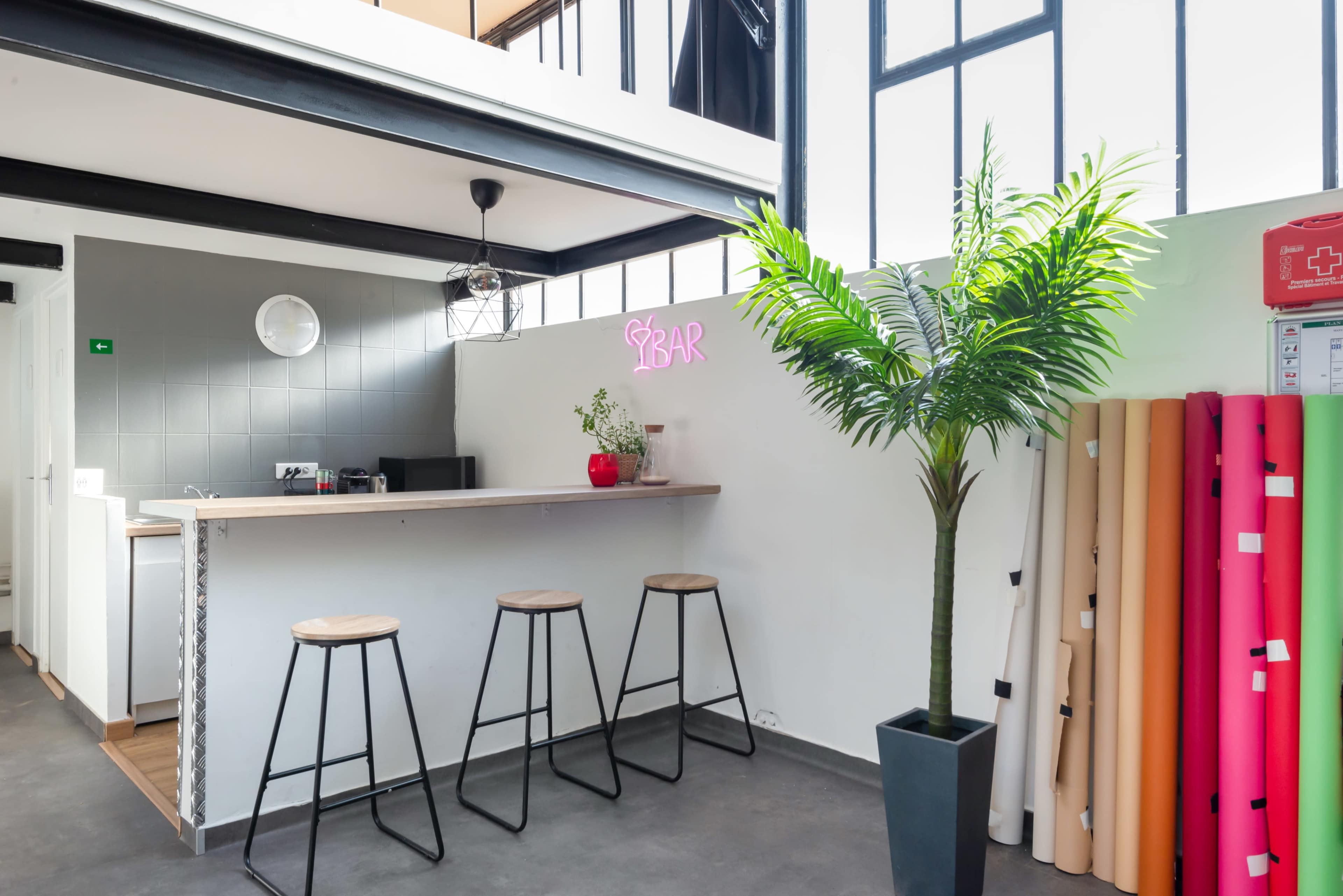 A modern kitchen space with a small bar area featuring three stools, a potted plant, and colorful rolls of material stacked against the wall.