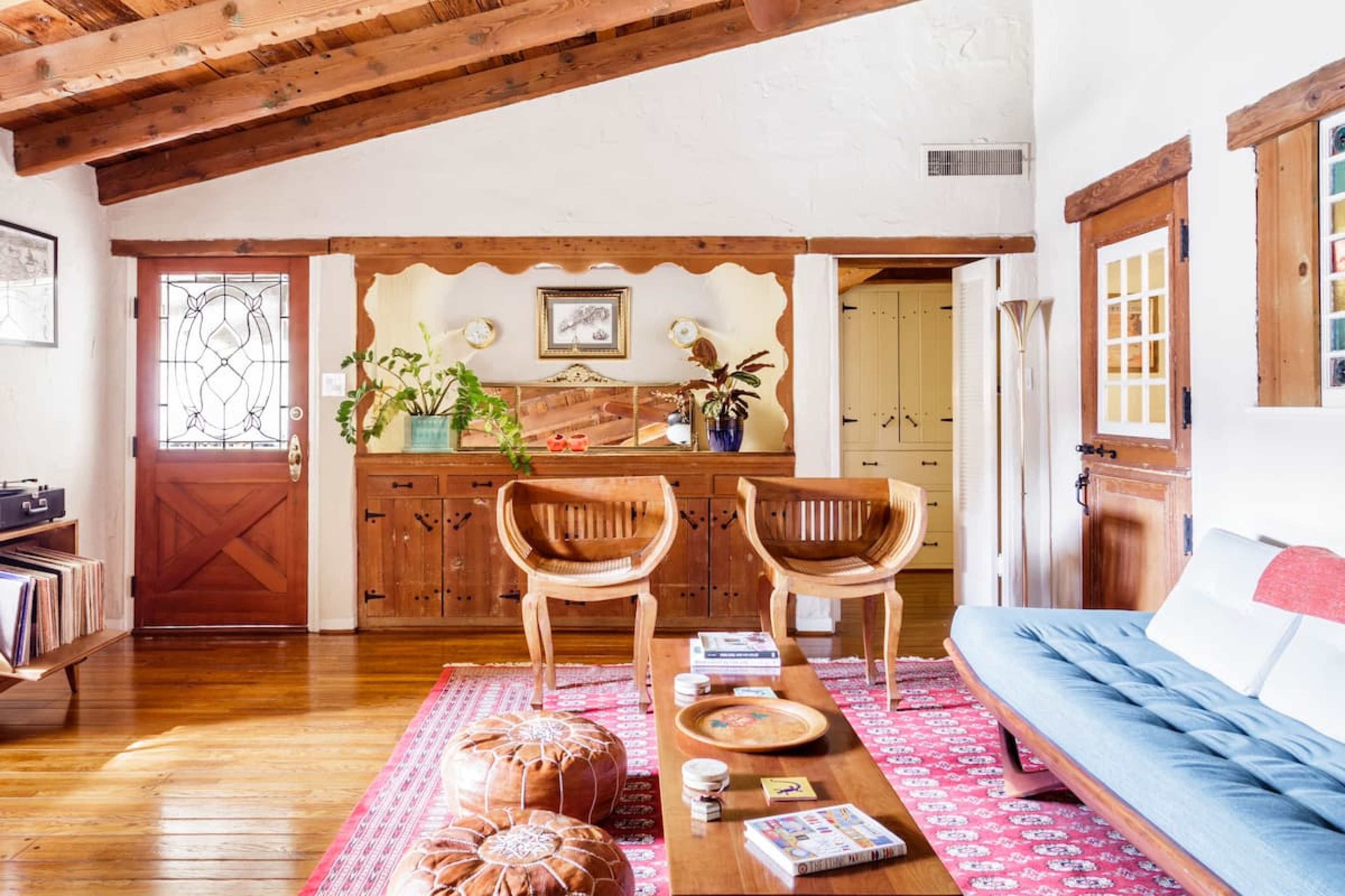 A cozy living room features wooden beams, a blue sofa, two wooden chairs, a coffee table with books, and a decorative entrance door.