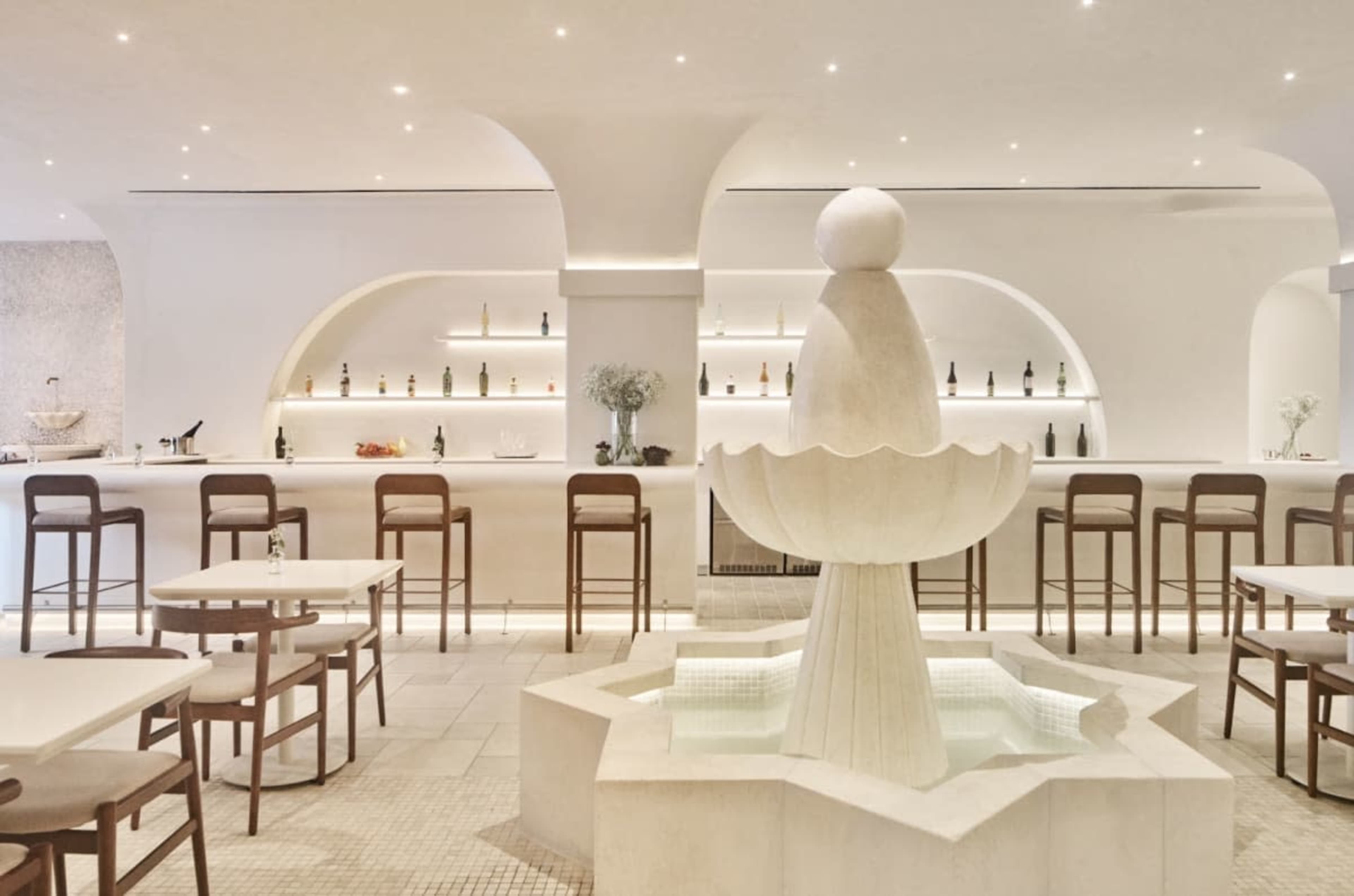 The image shows a modern restaurant with a white fountain at its center, surrounded by light-colored seating and a bar lined with bottles.