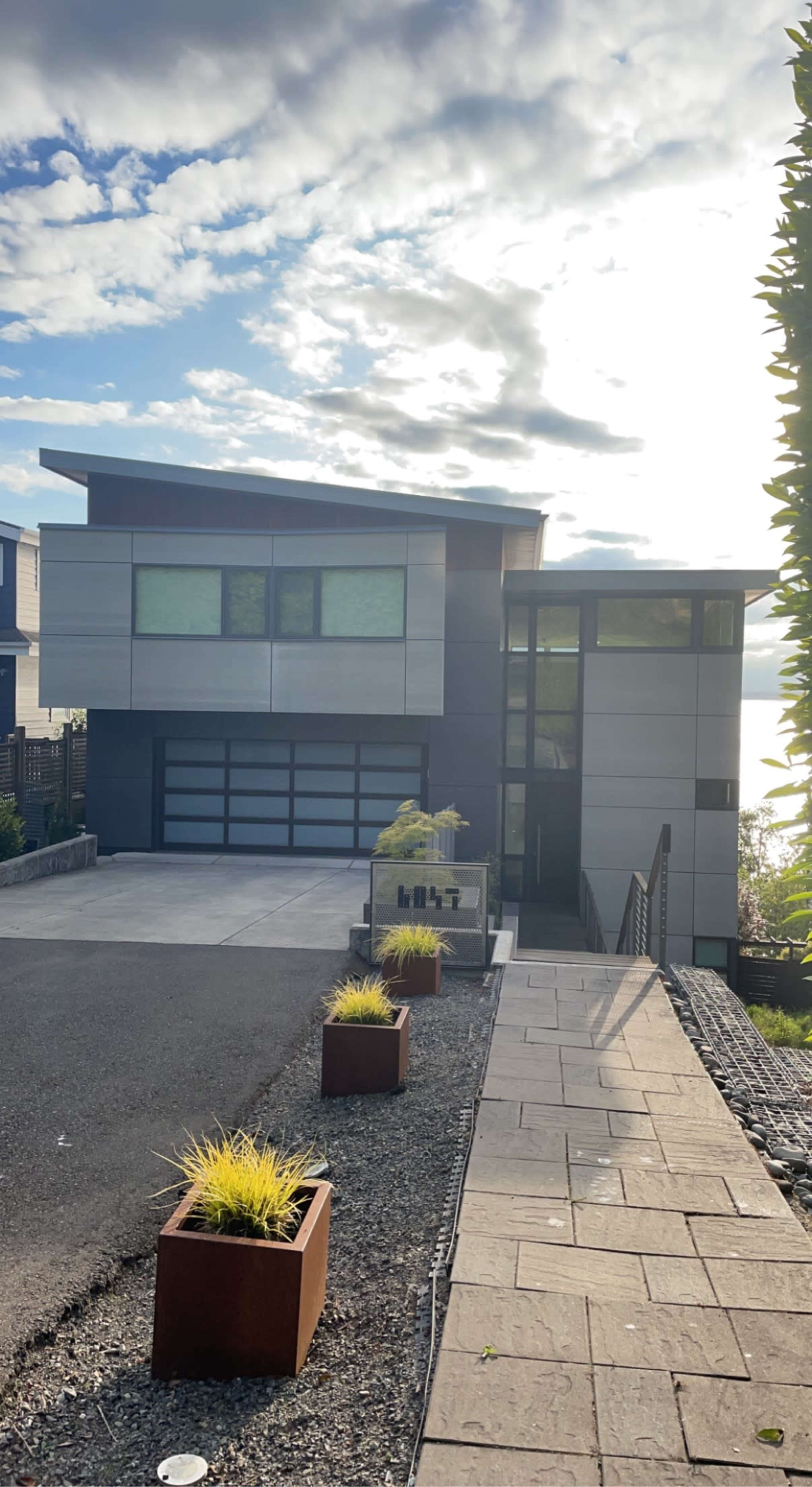 A modern two-story house with a rectangular design and large windows is situated at the end of a paved driveway, surrounded by landscaped planters.