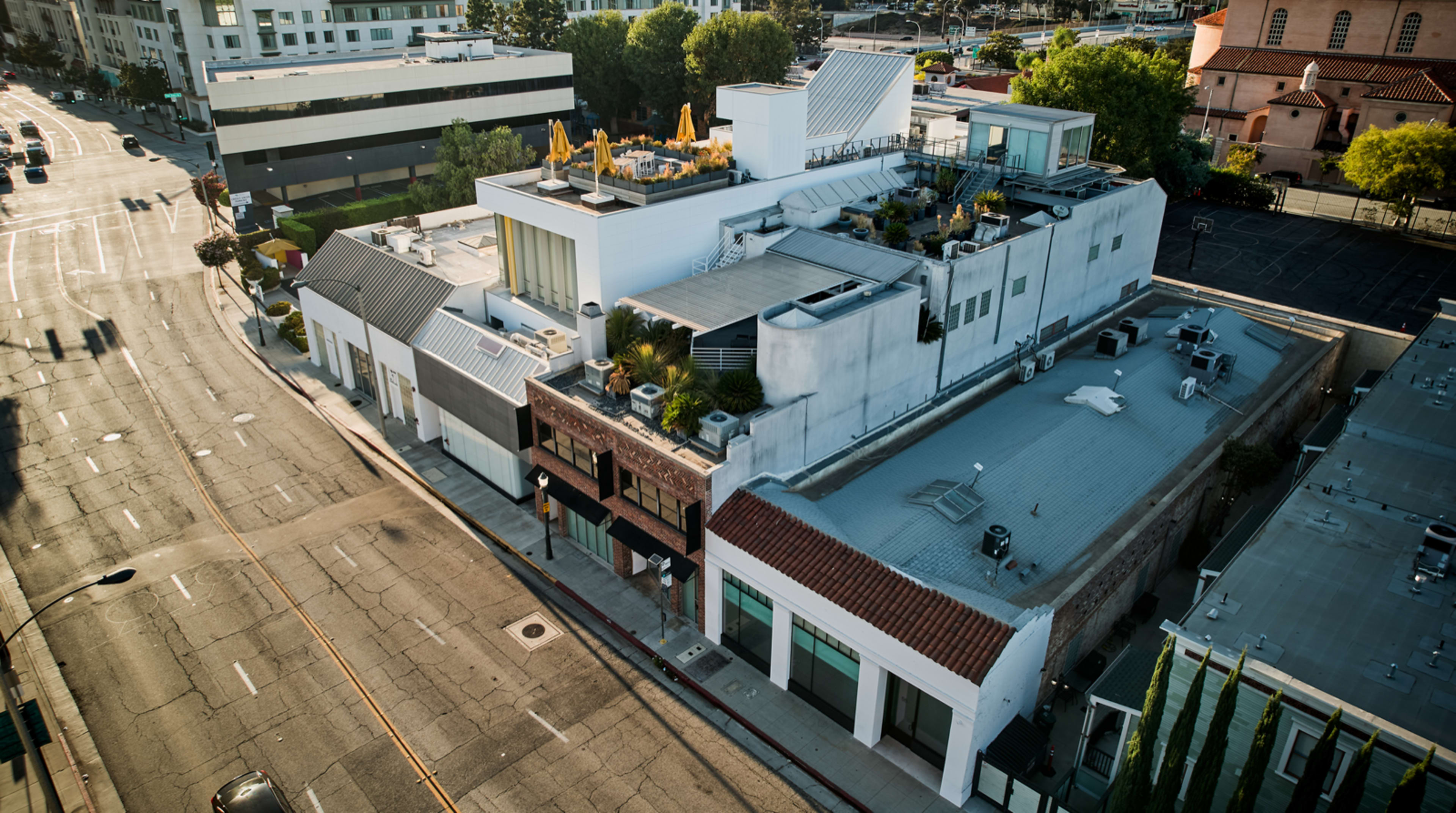The image shows a modern building complex with a rooftop terrace, situated at the corner of a city street, surrounded by parking areas and trees.