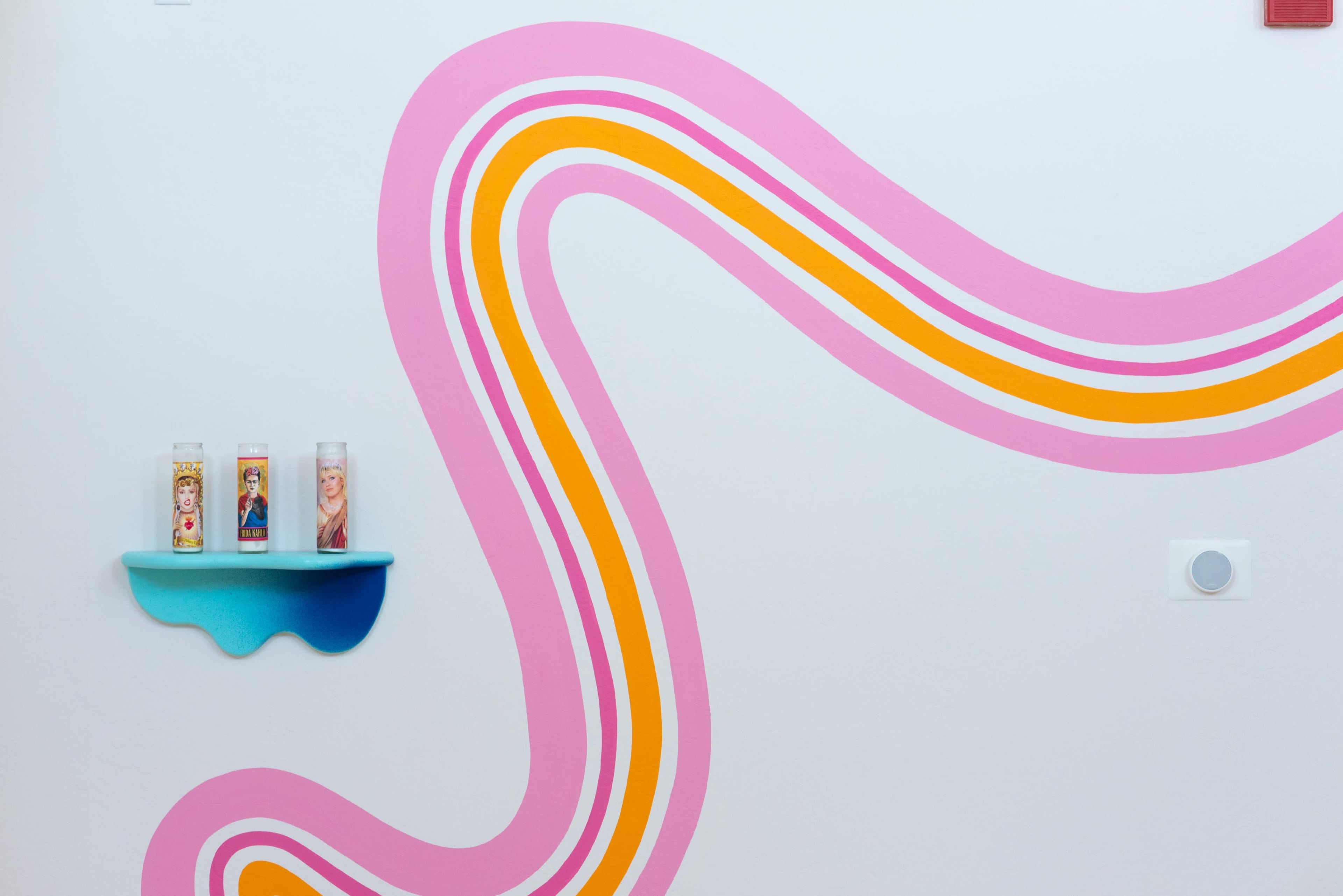 The image features a wall adorned with colorful curved stripes and a small shelf holding three candles.