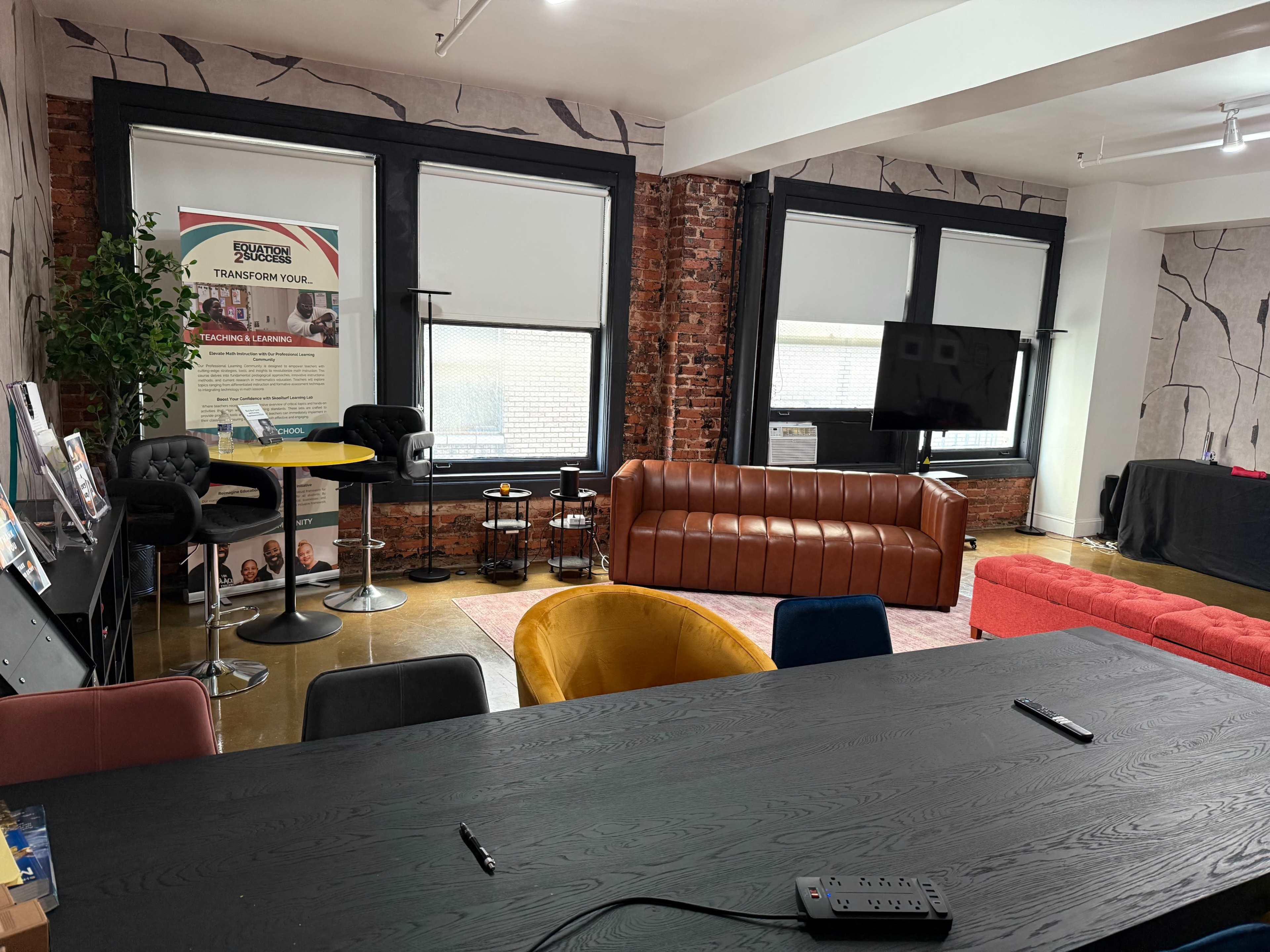The image shows a modern office space with a brown leather sofa, a yellow chair, and a long black table, along with various decor elements and a large screen on the wall.