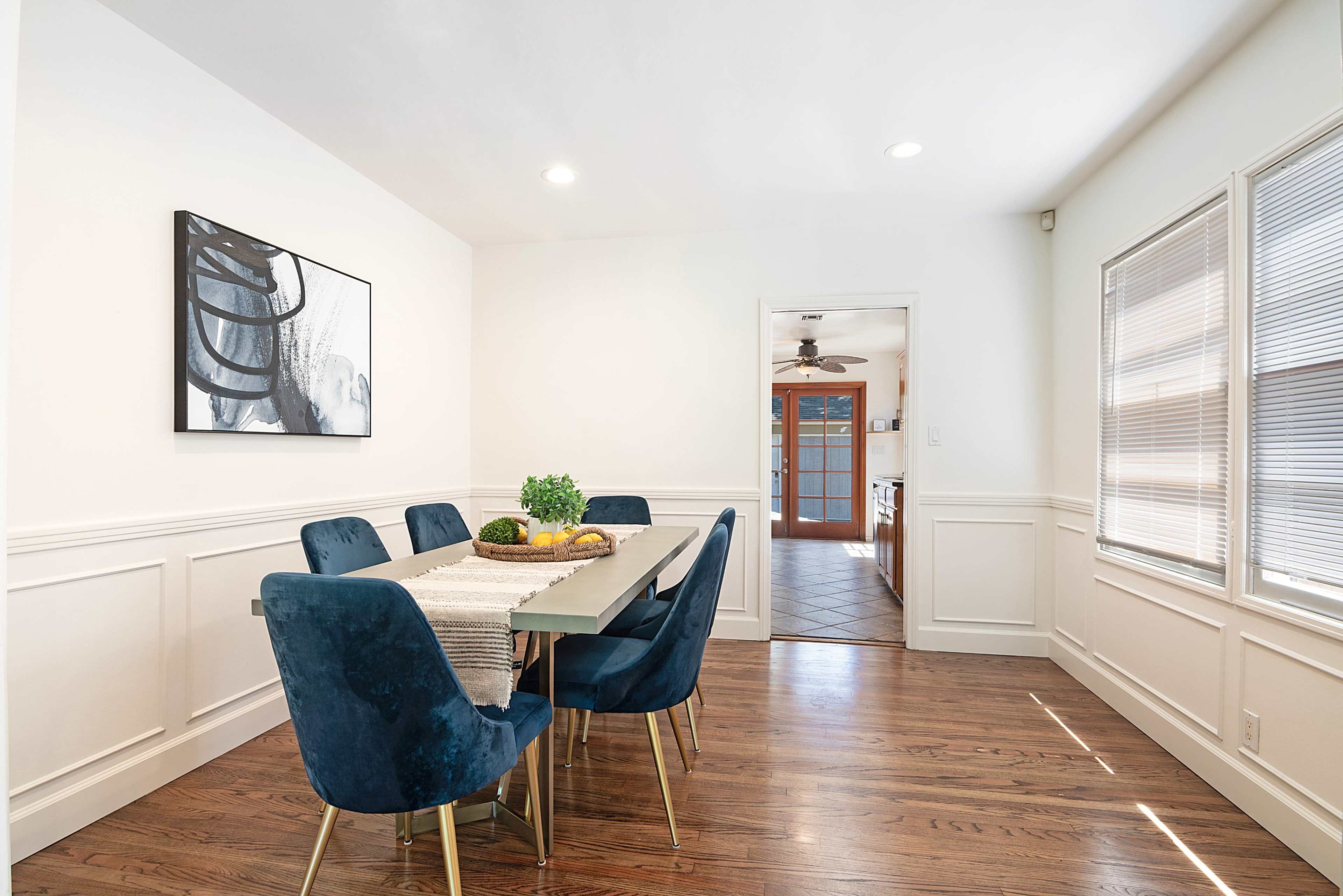 A bright dining room features a rectangular table surrounded by six blue chairs, with a door leading to a kitchen in the background.