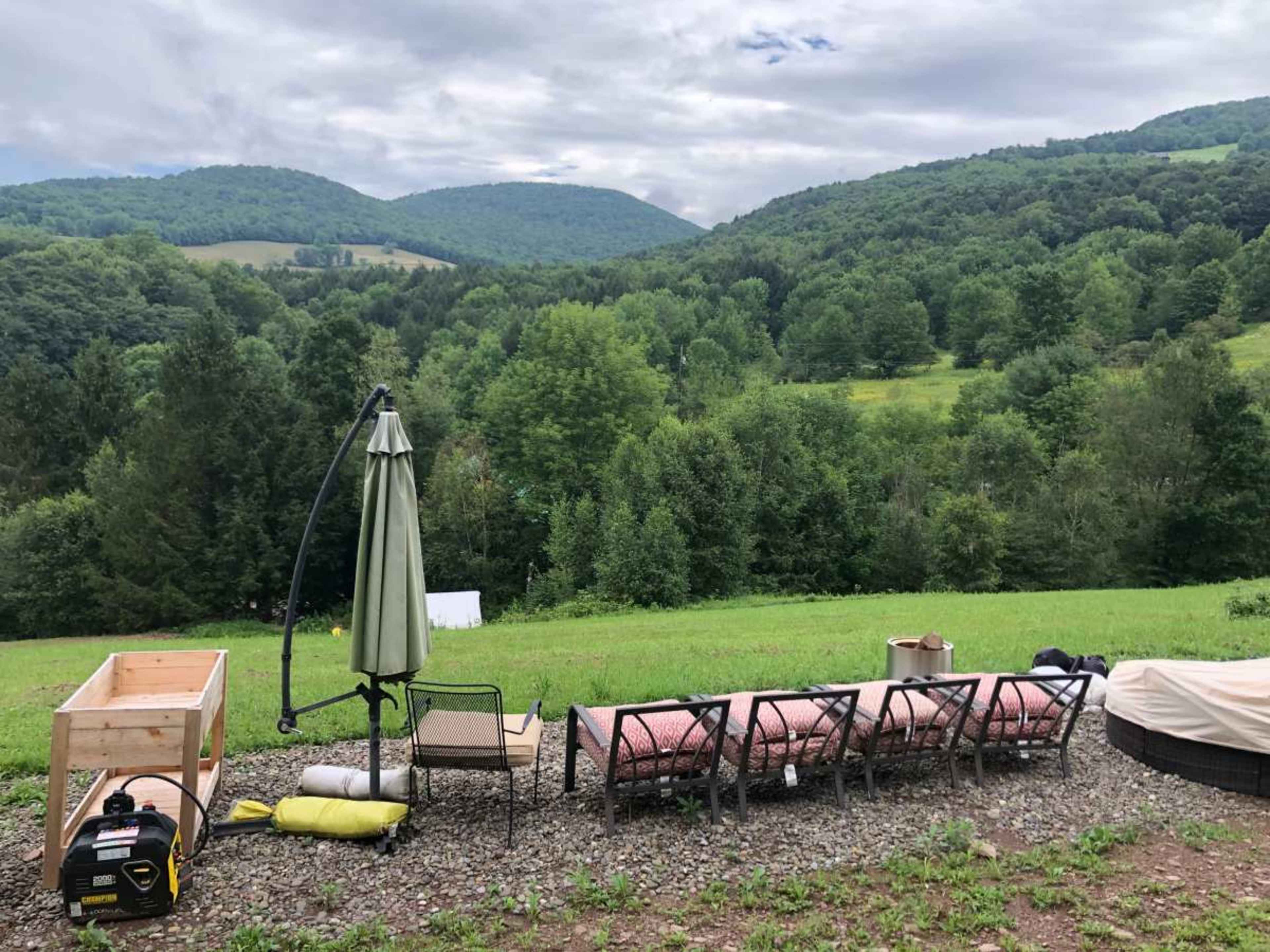 A row of outdoor chairs and a table overlook a lush, green valley surrounded by mountains under a cloudy sky.