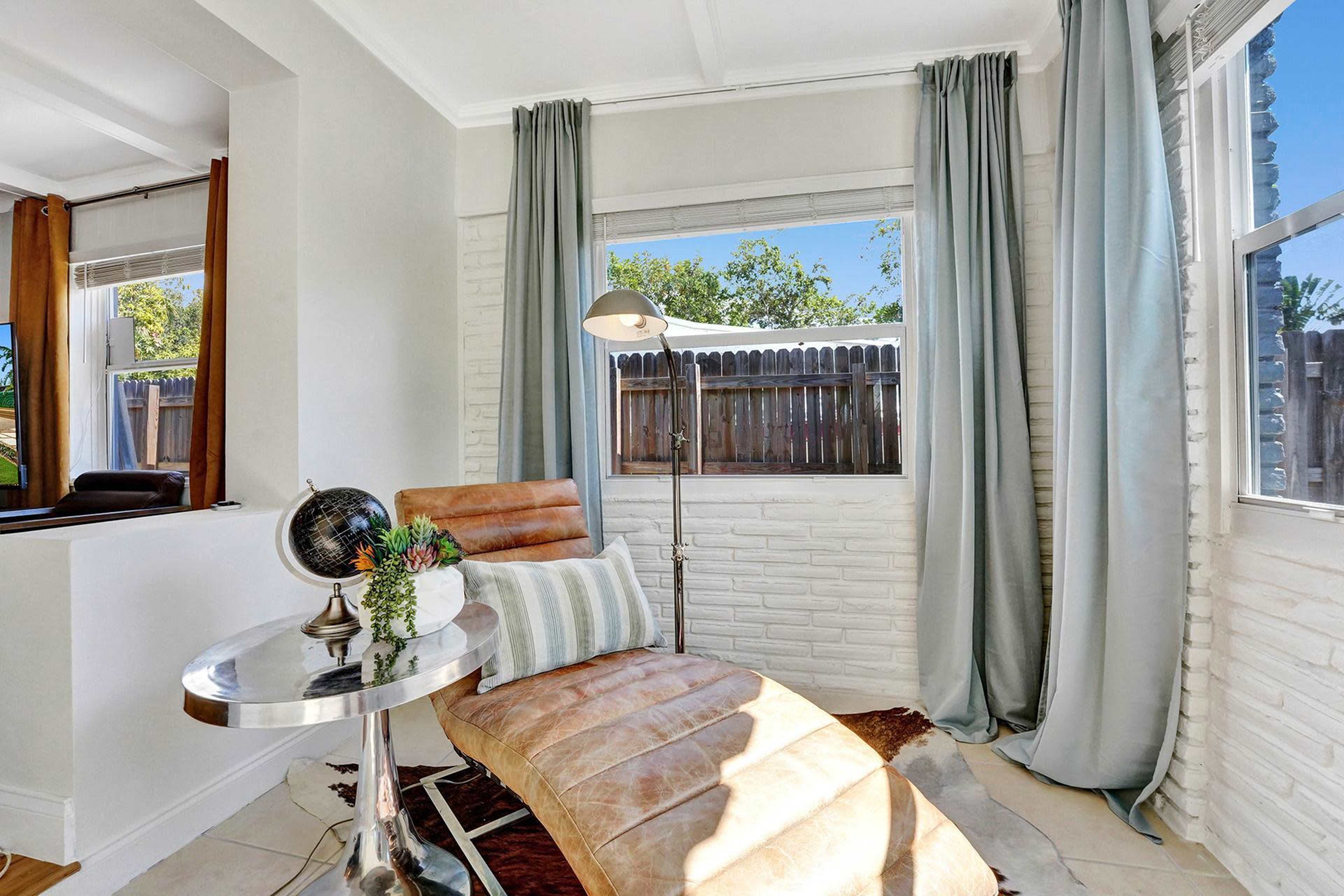 A cozy reading nook features a leather chair, a small metallic table with potted plants, and large windows with light curtains.
