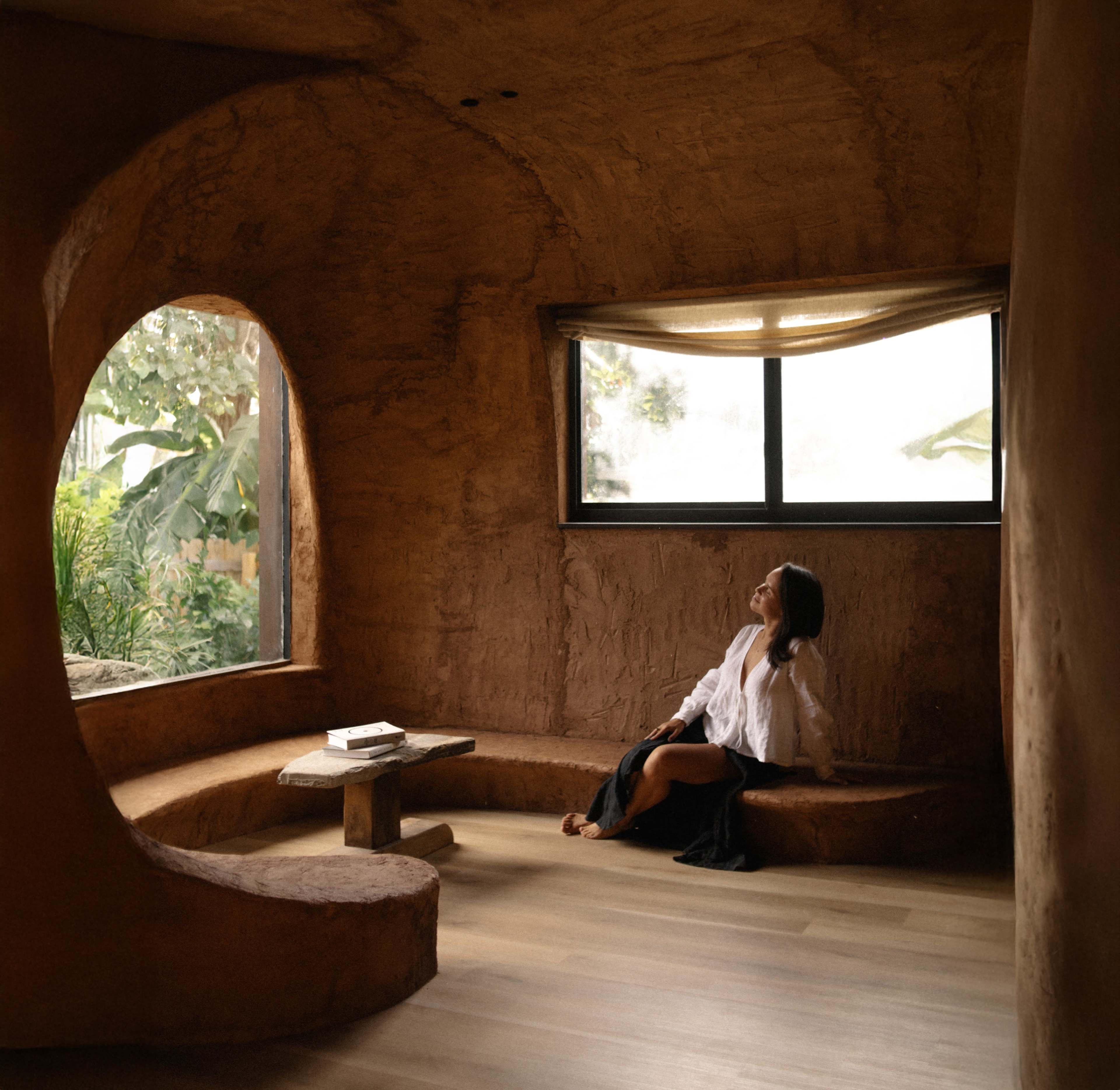 A woman sits in a curved, earthy-toned room with large windows that provide a view of greenery outside.