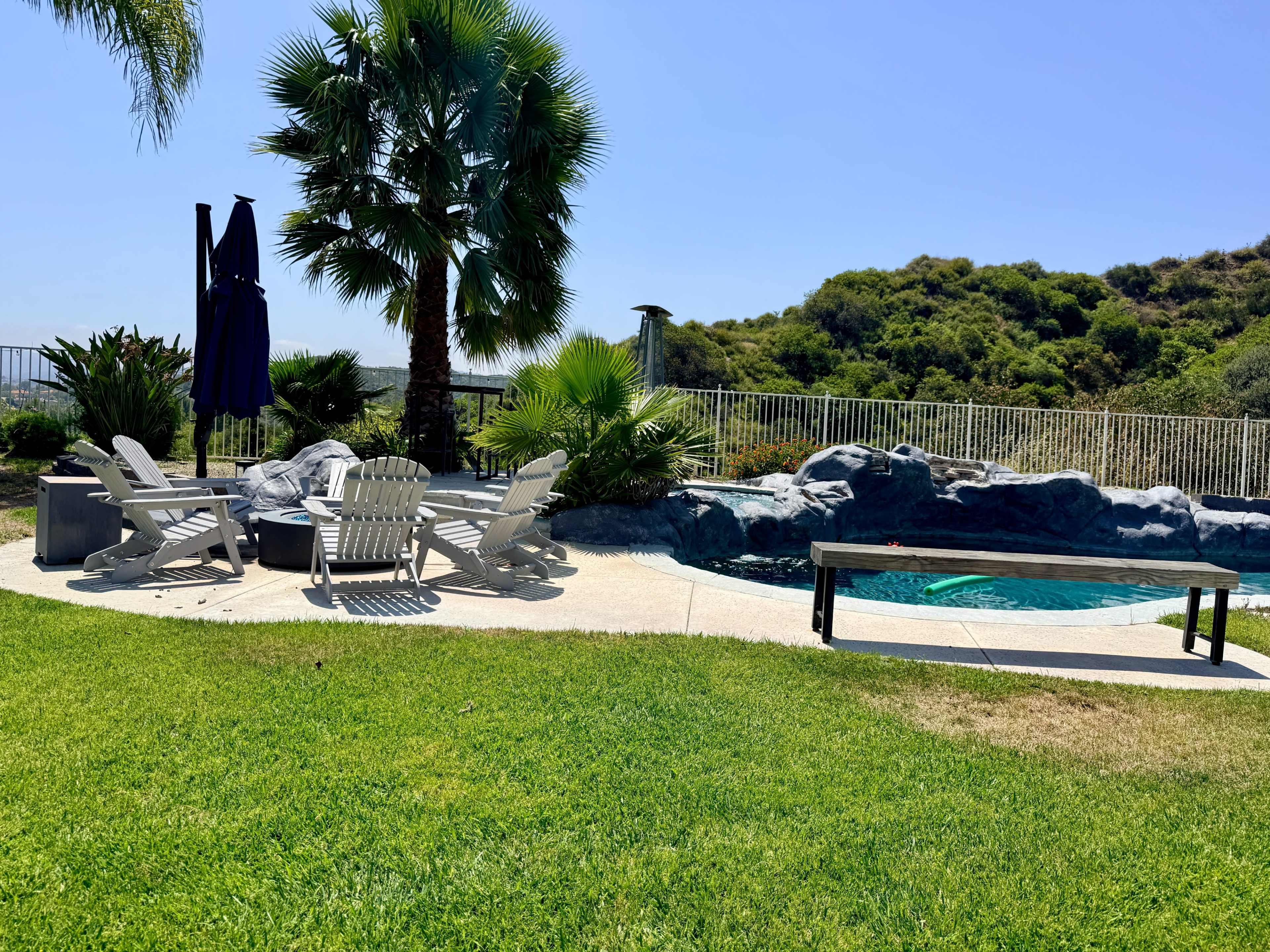 The scene features a poolside area with white lounge chairs and a table near a rock formation and lush greenery.