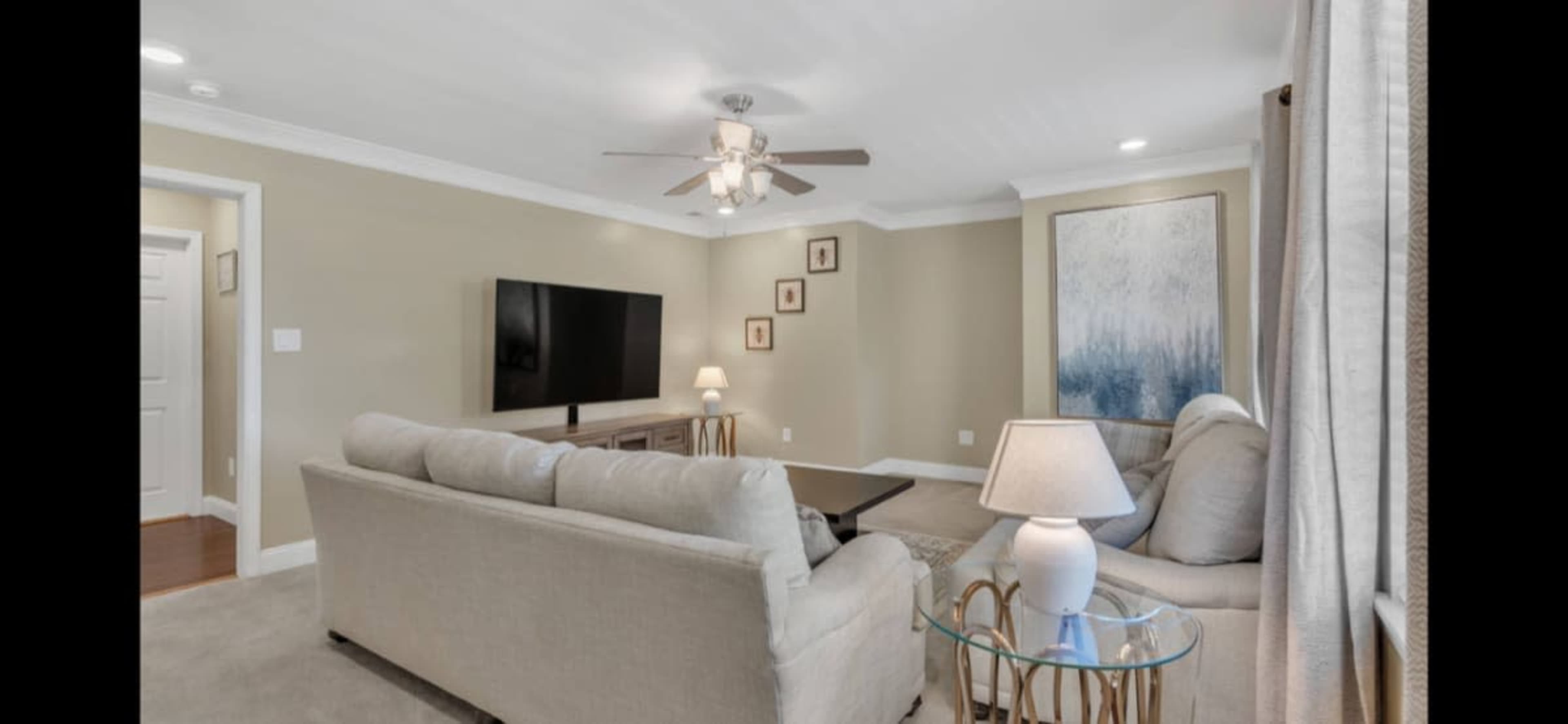 A living room with a light-colored sofa, a television mounted on the wall, and a lamp on a side table, complemented by decor and wall art.