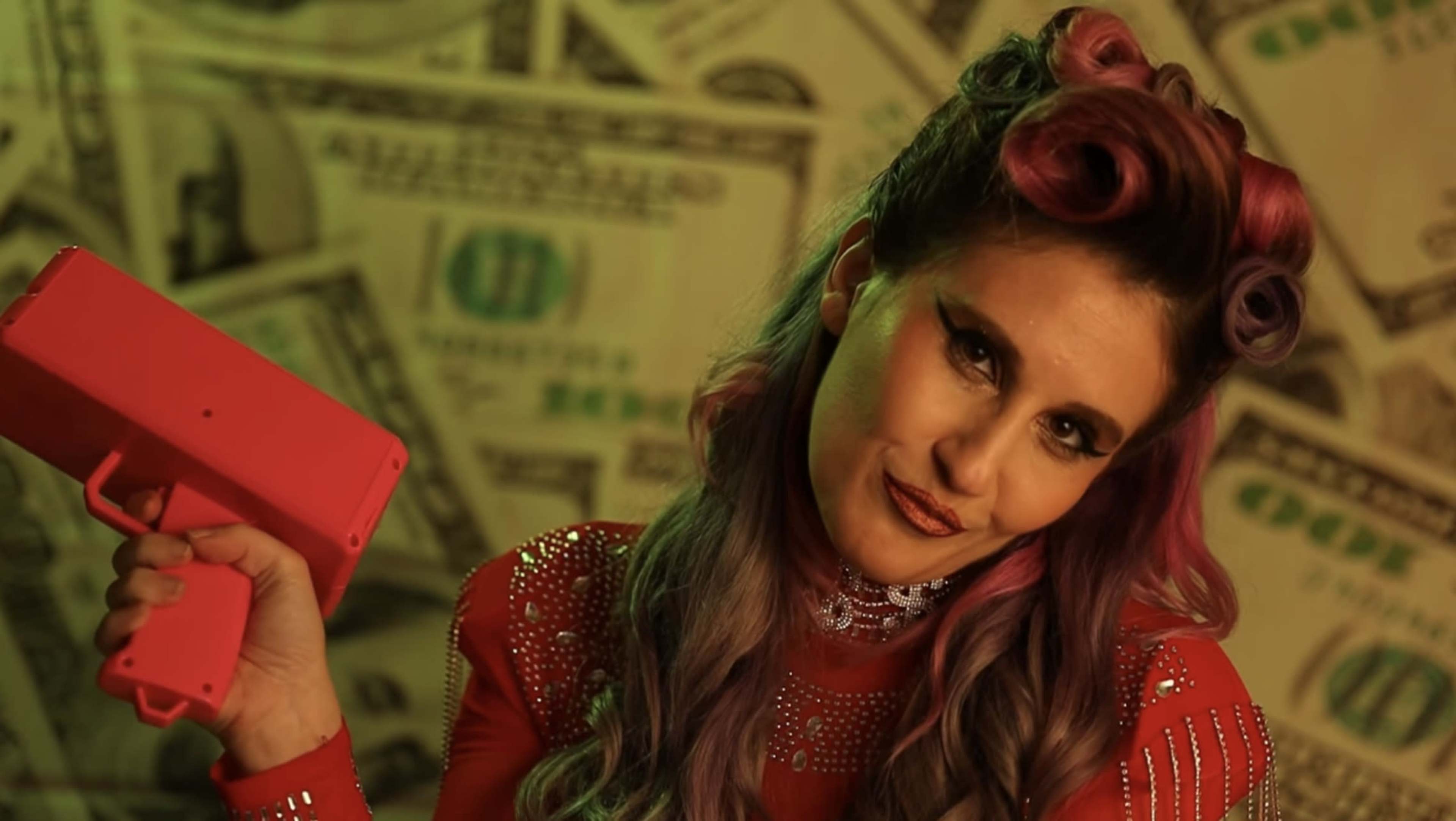 A woman with pink curls holds a toy gun in front of a backdrop covered in images of money.