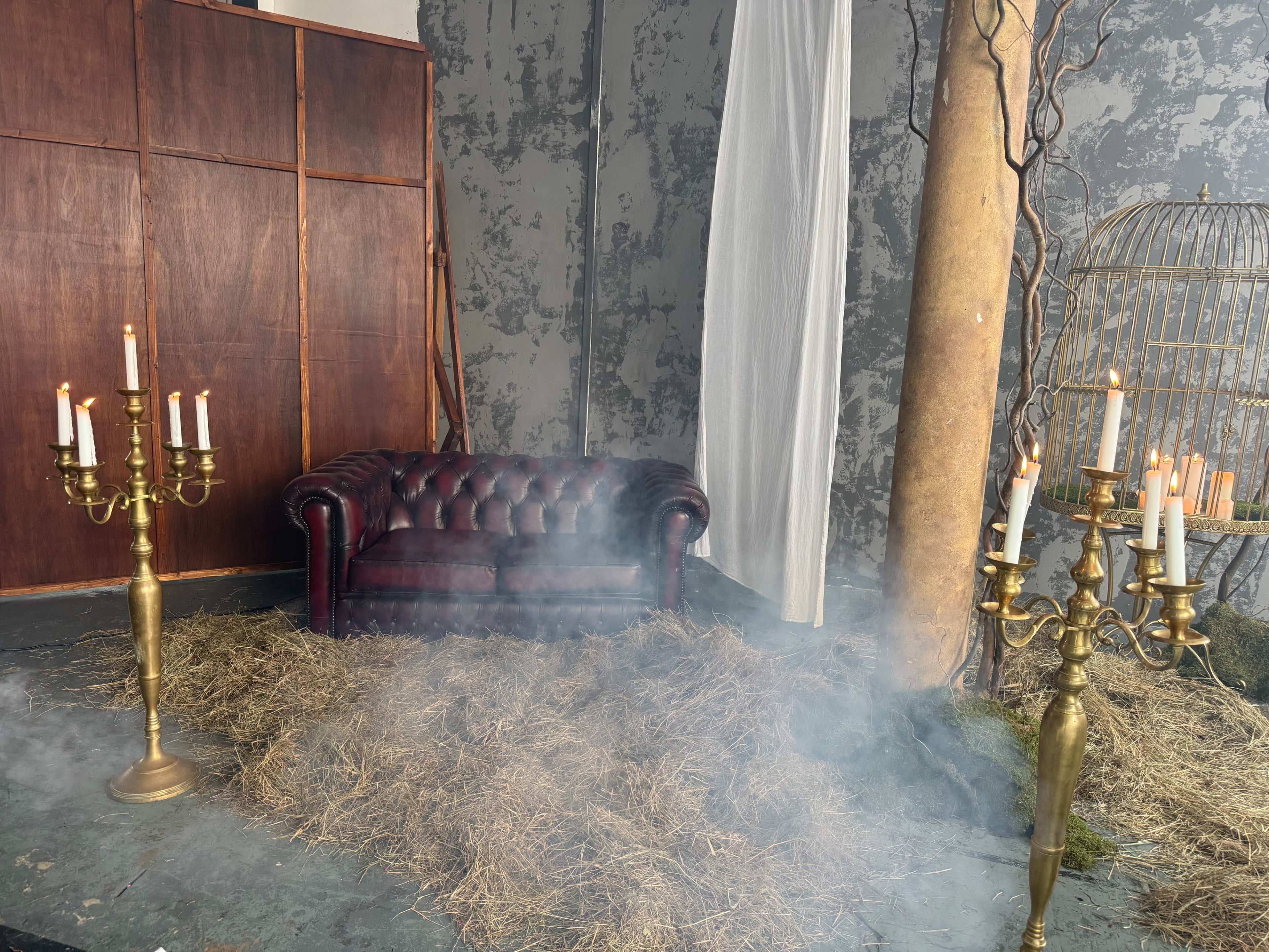 The scene features a brown velvet couch surrounded by two ornate candelabra and a birdcage, with hay scattered on the floor and smoke gently filling the area.