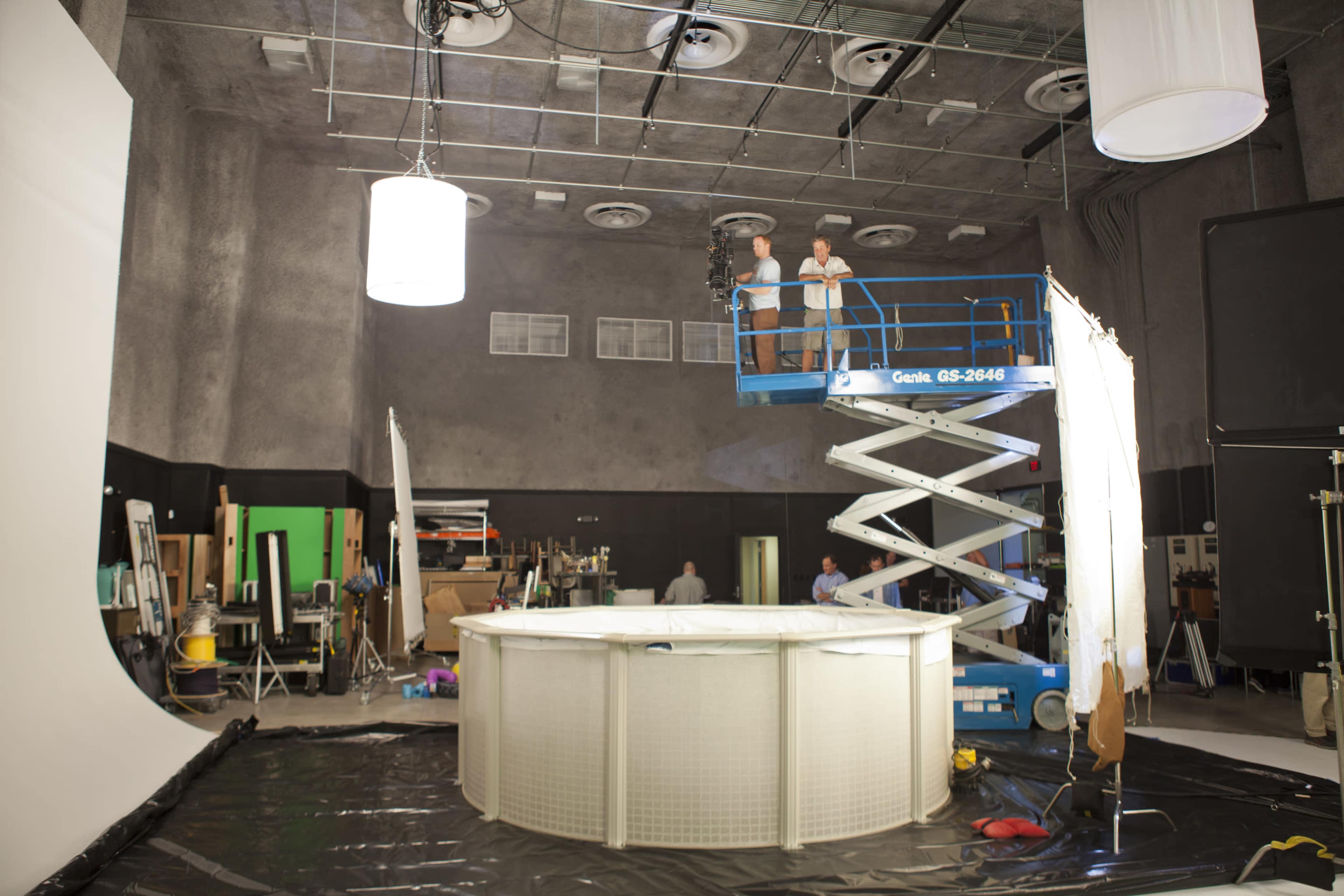 A crew member operates a scissor lift above a circular set surrounded by various film equipment and backdrops in a studio.