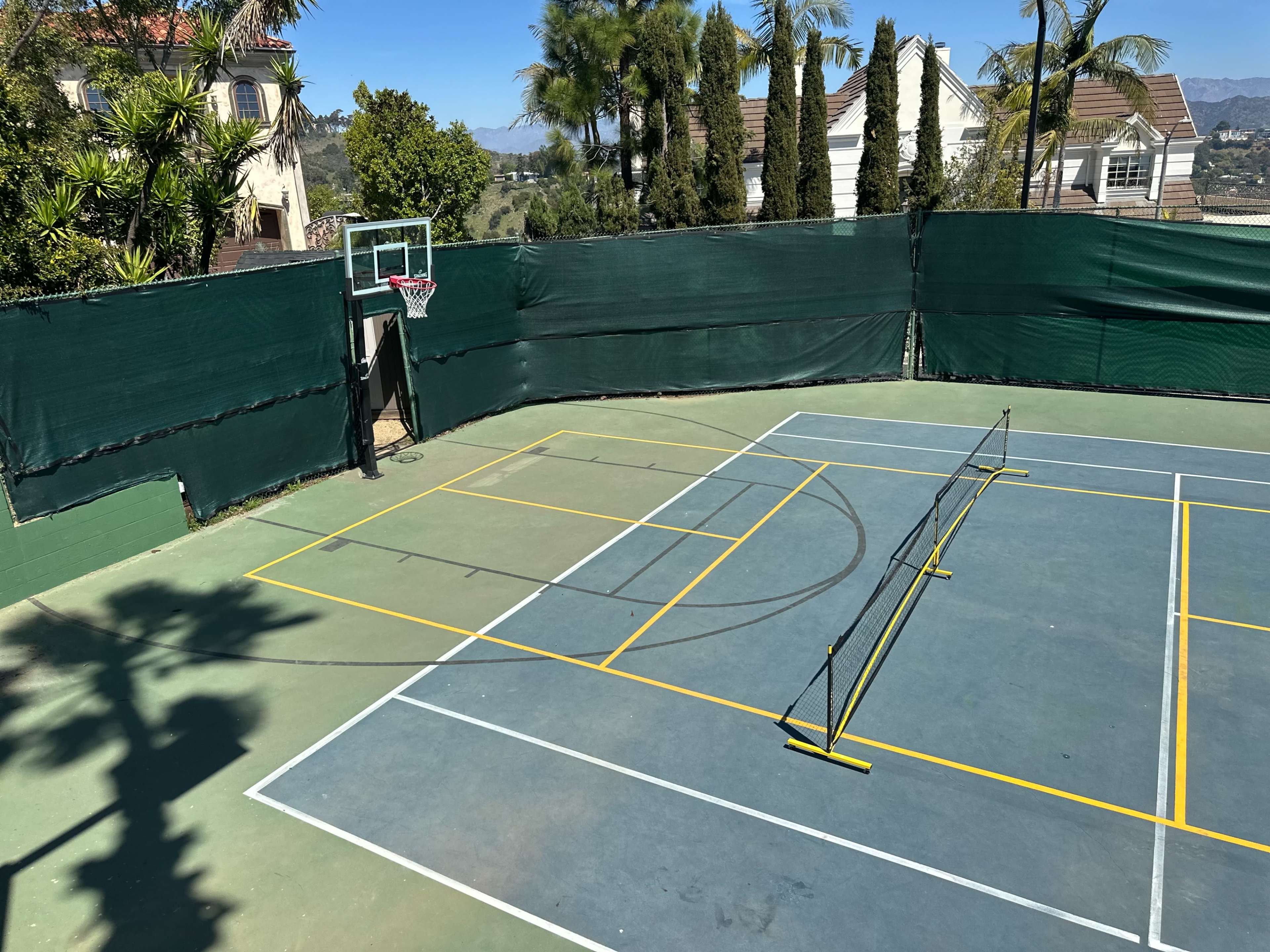 The image shows a secluded sports court, comprising a half basketball court and a netted area for tennis, surrounded by green fencing and palm trees.