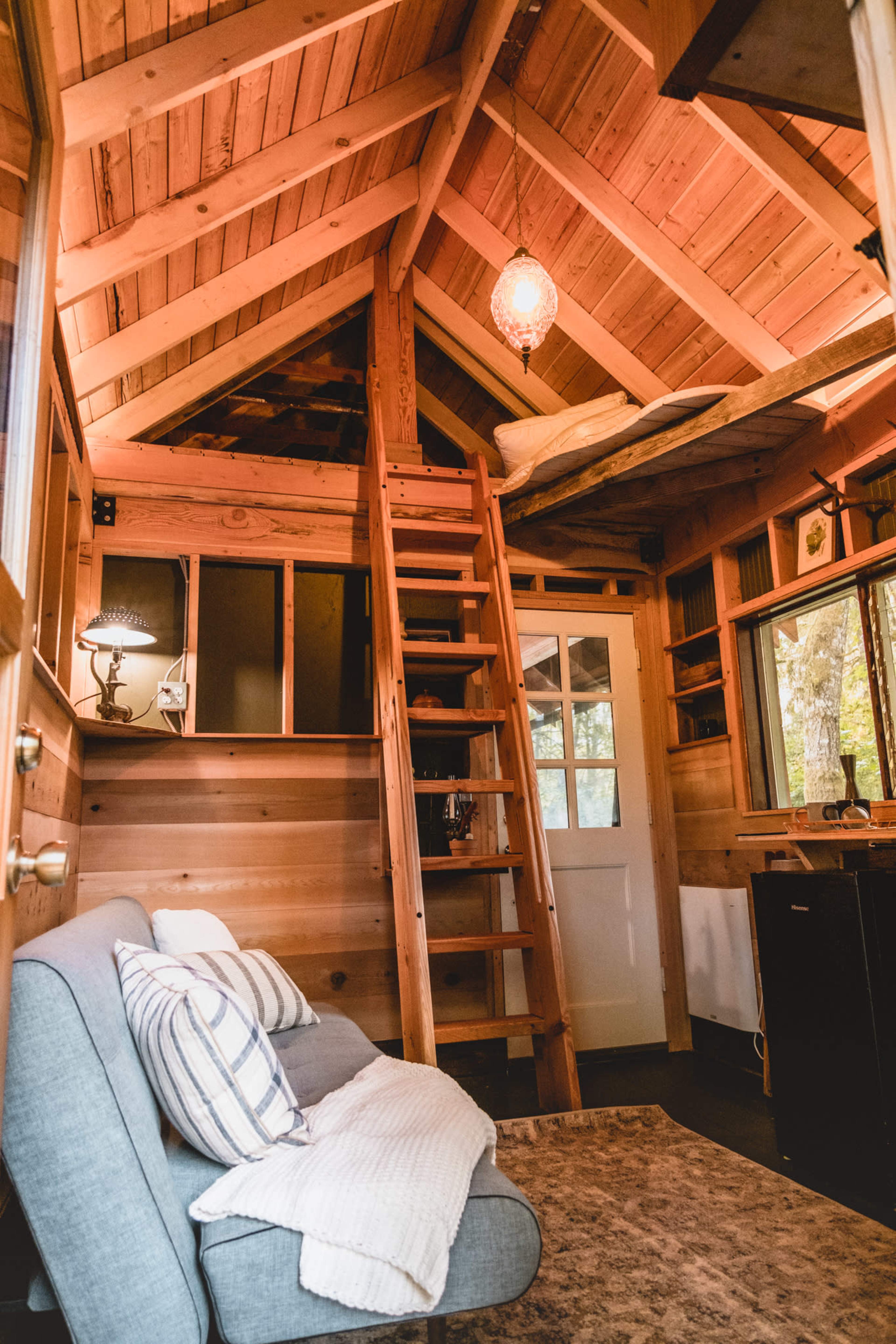 The interior of a wooden tiny house features a cozy sofa, a ladder leading to a loft, and warm wooden walls with a pendant light.