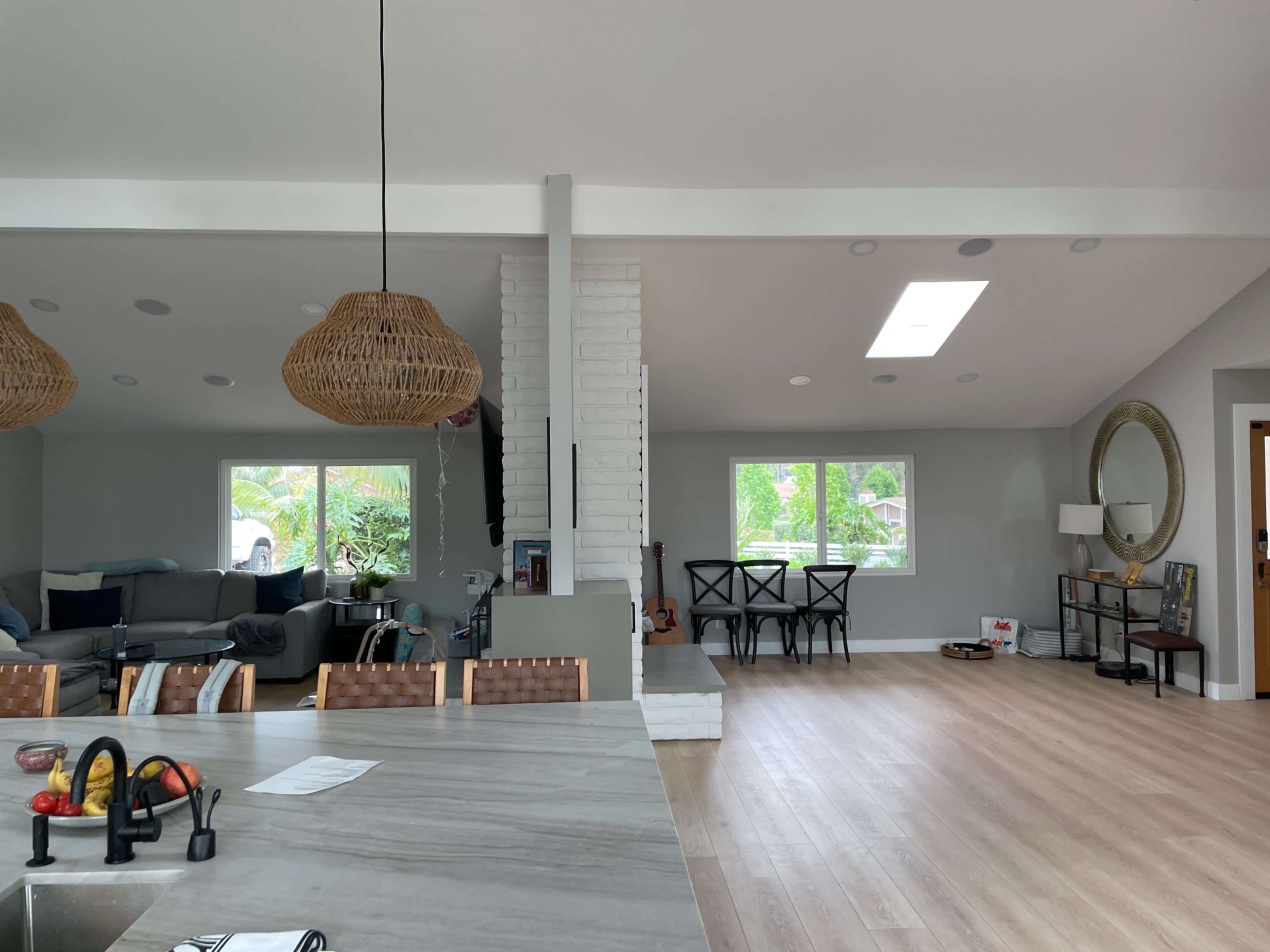 The image shows an open-concept living area with a kitchen on the left and a seating area to the right, featuring large windows and wooden flooring.