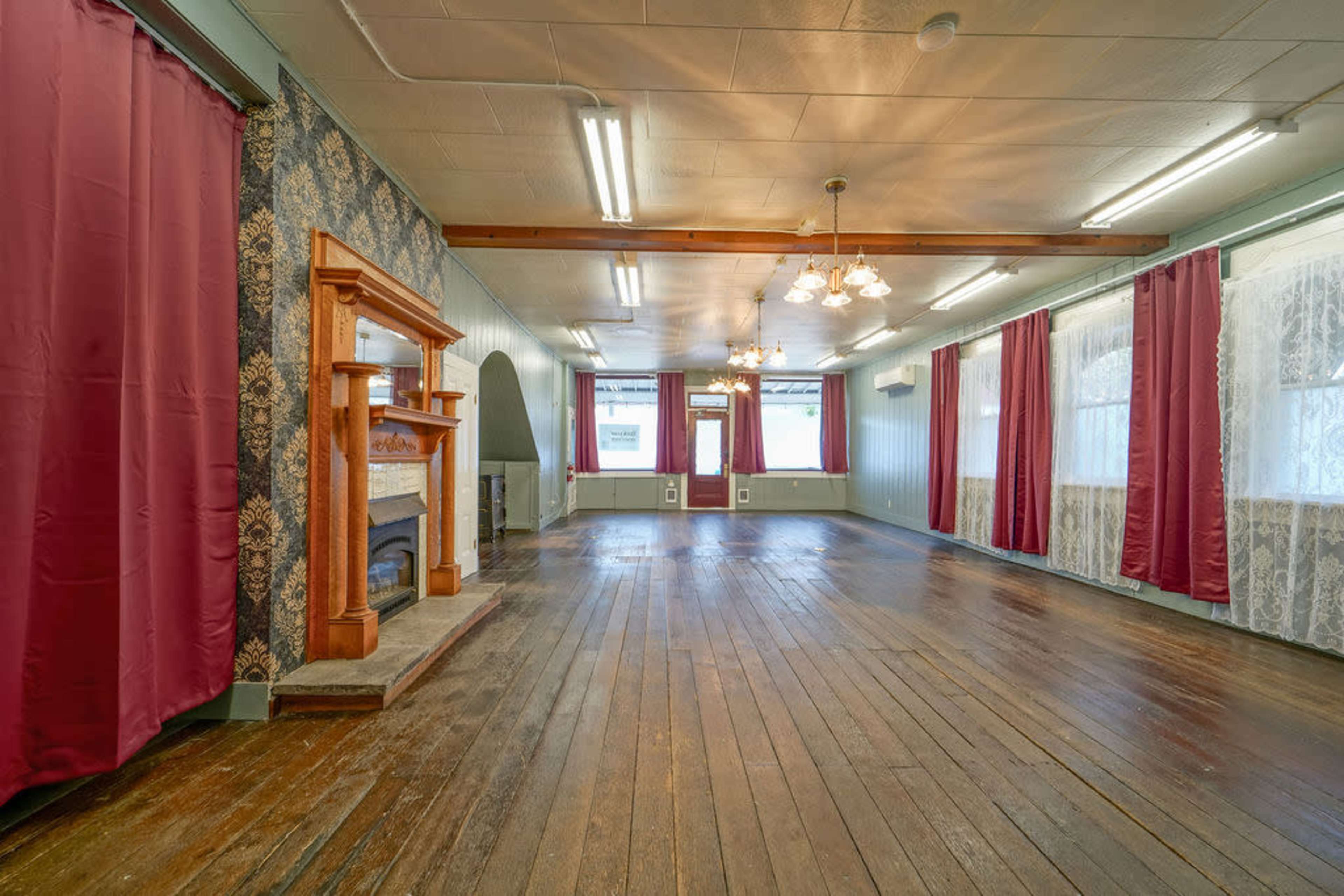 The image shows an empty room with hardwood floors, decorated with red curtains, a vintage fireplace, and a patterned wall.
