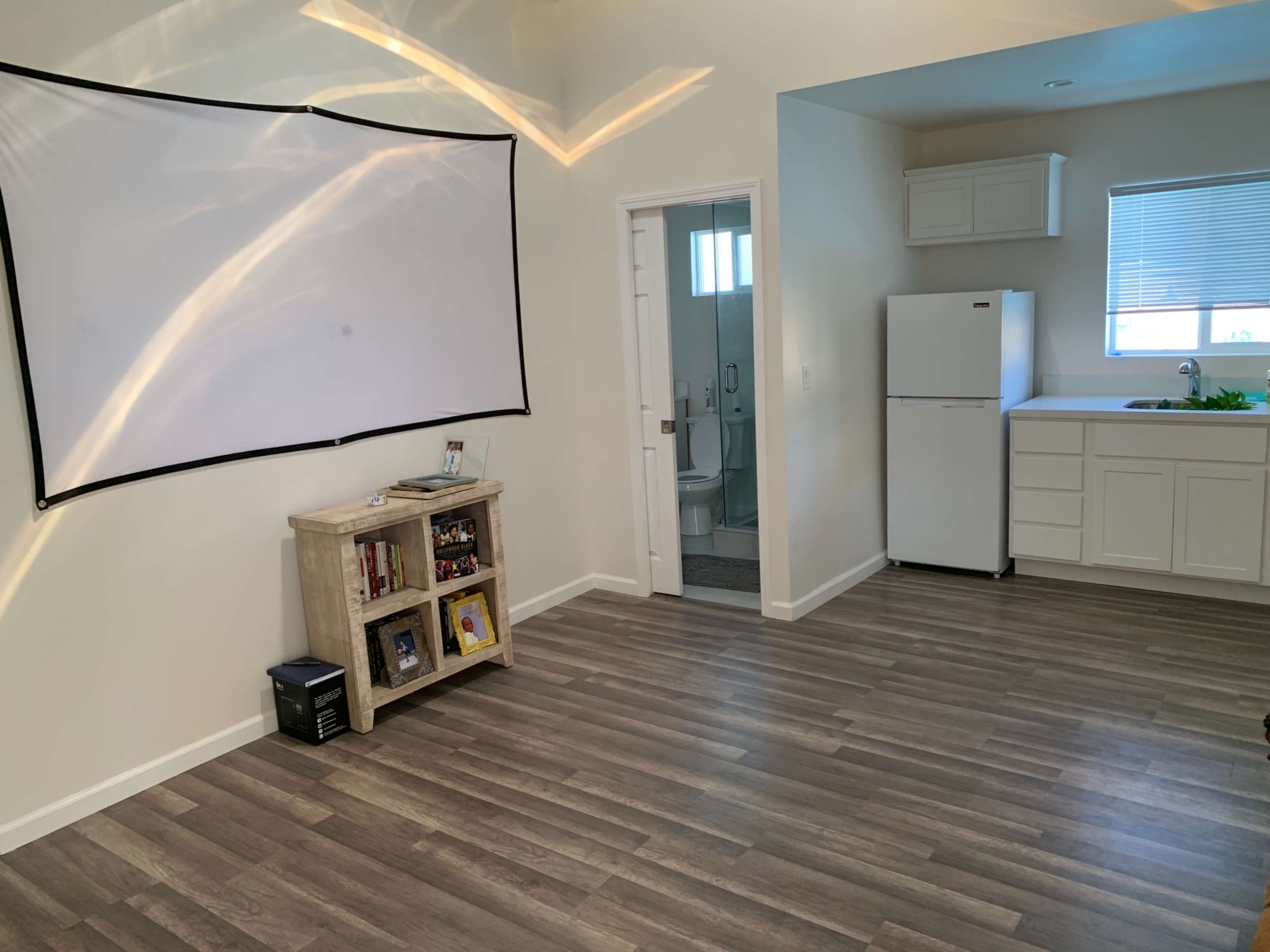 The image shows a room featuring a large projection screen, a bookshelf, a refrigerator, and a small kitchen area, with a bathroom visible through an open door.