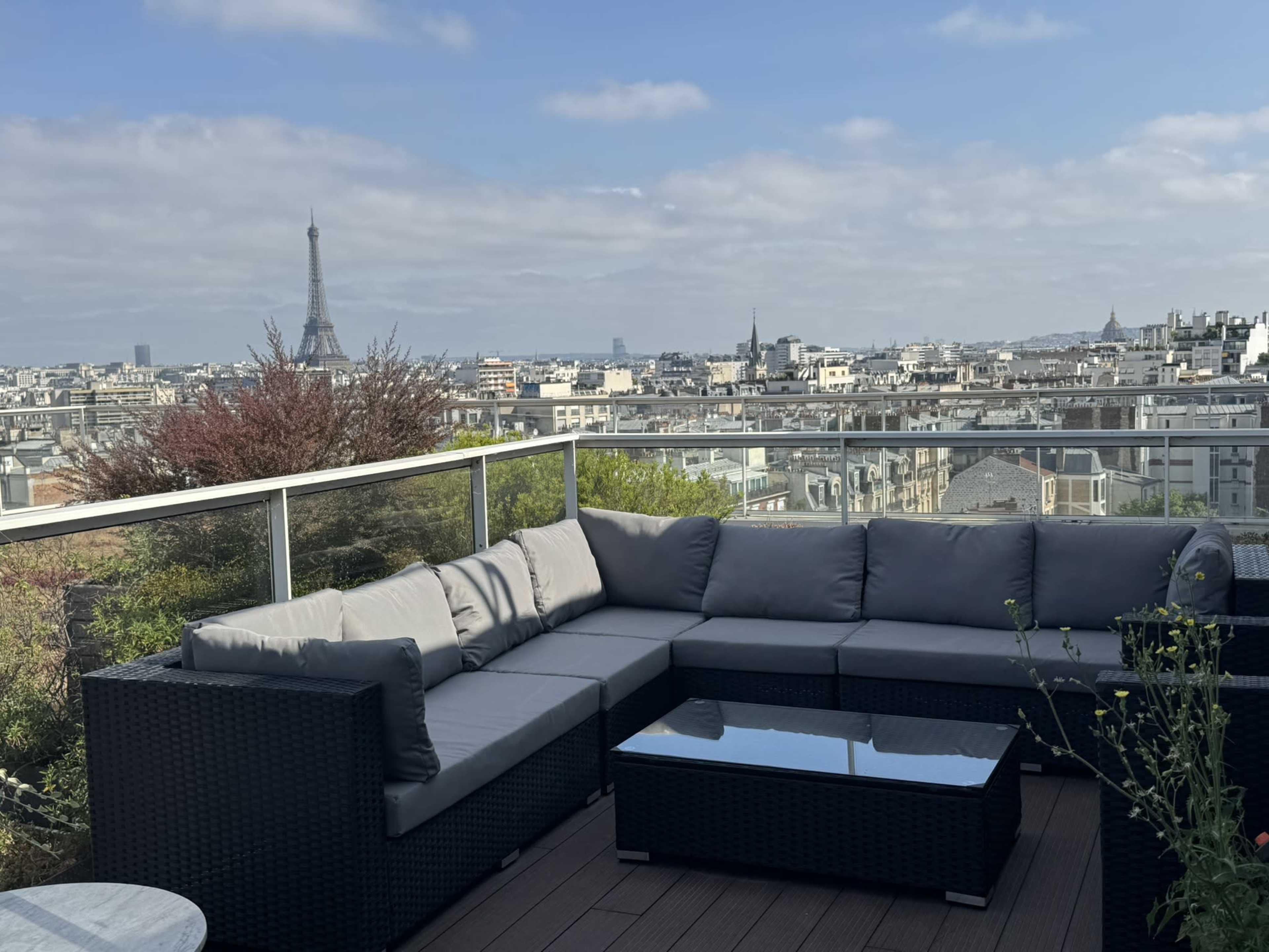 A rooftop terrace features a gray sectional sofa and a glass table, overlooking a cityscape with the Eiffel Tower in the distance.