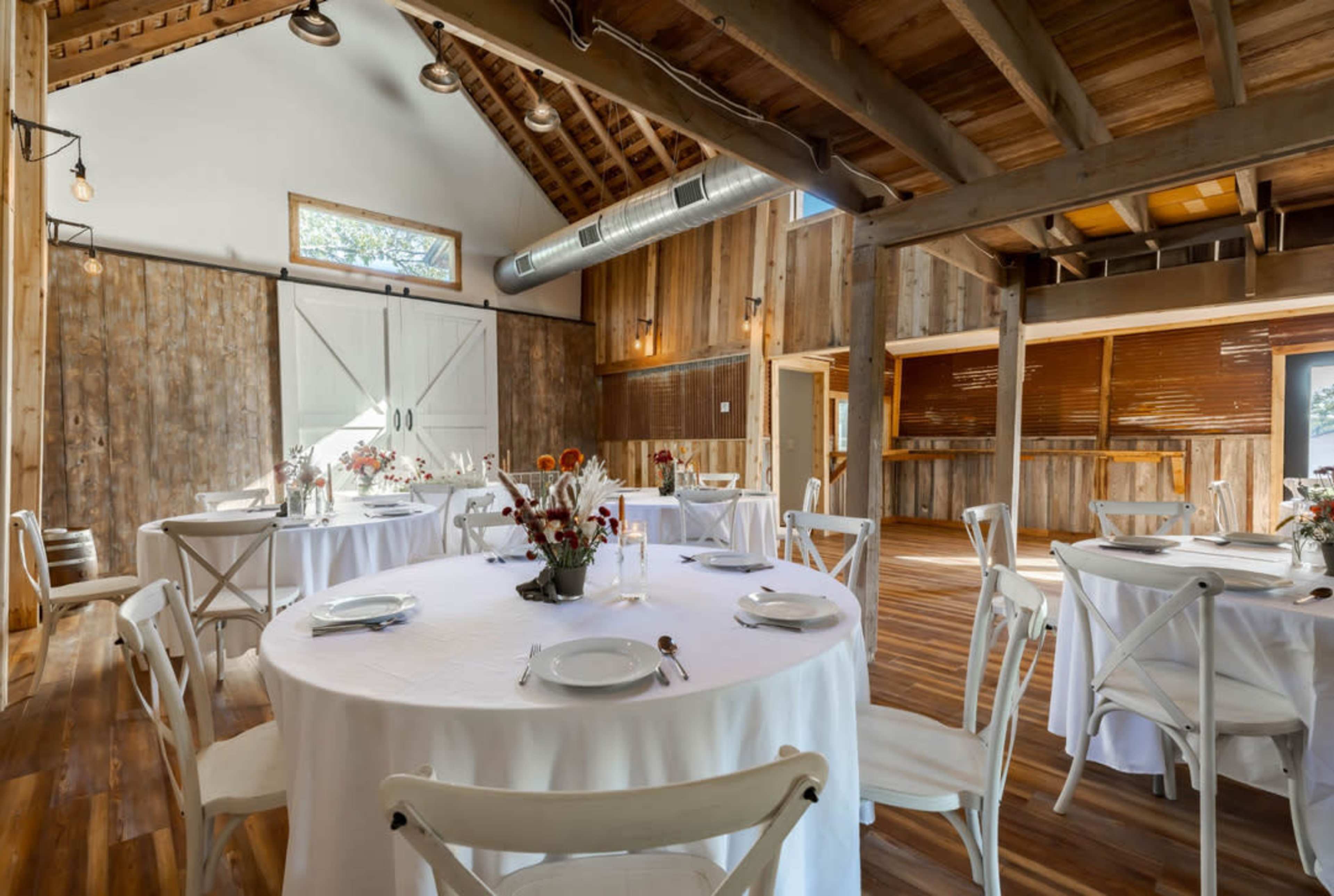 A rustic venue features round tables set with white tablecloths and a floral centerpiece, illuminated by natural light through large windows.