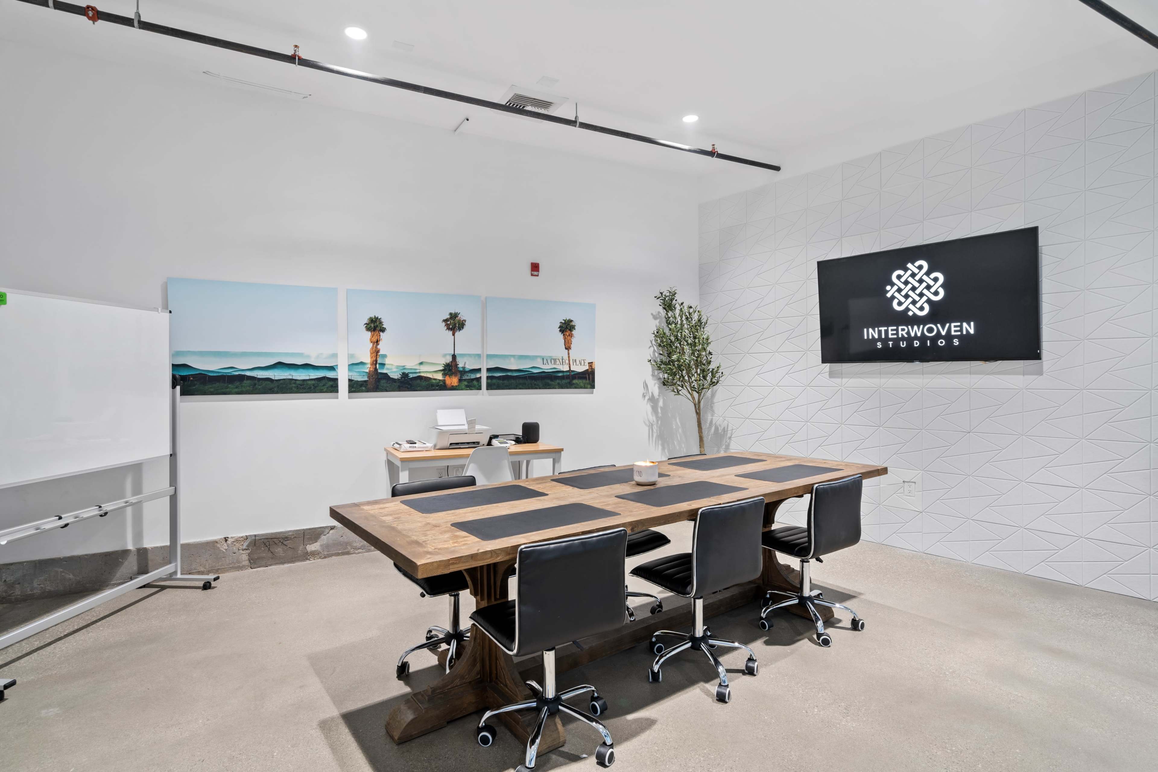 A modern conference room featuring a large wooden table with black chairs, a whiteboard, and artwork on the walls, with a logo displayed on a screen.