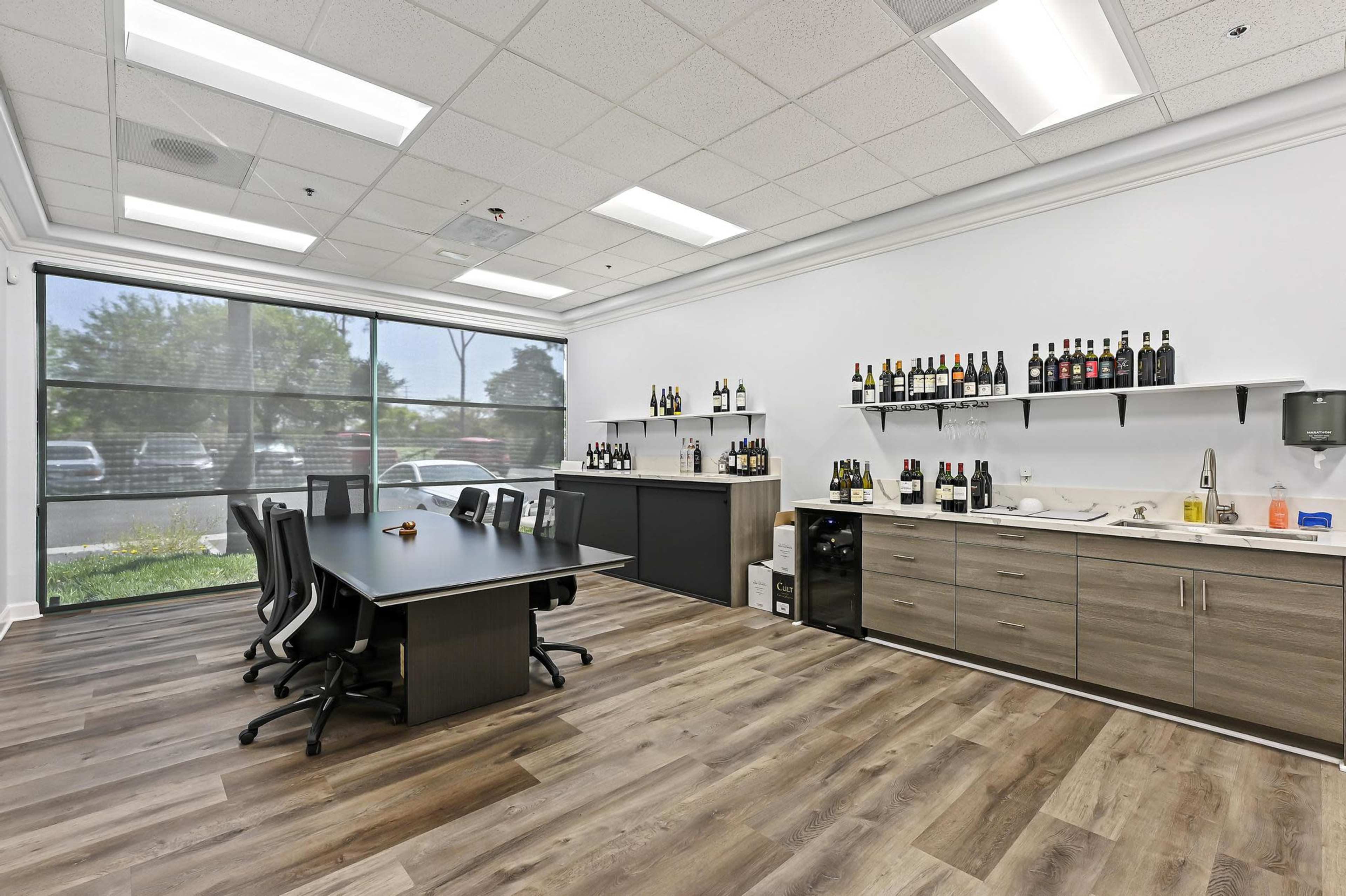 A modern office space with a large table, black chairs, a counter displaying various bottles, and a large window providing natural light.