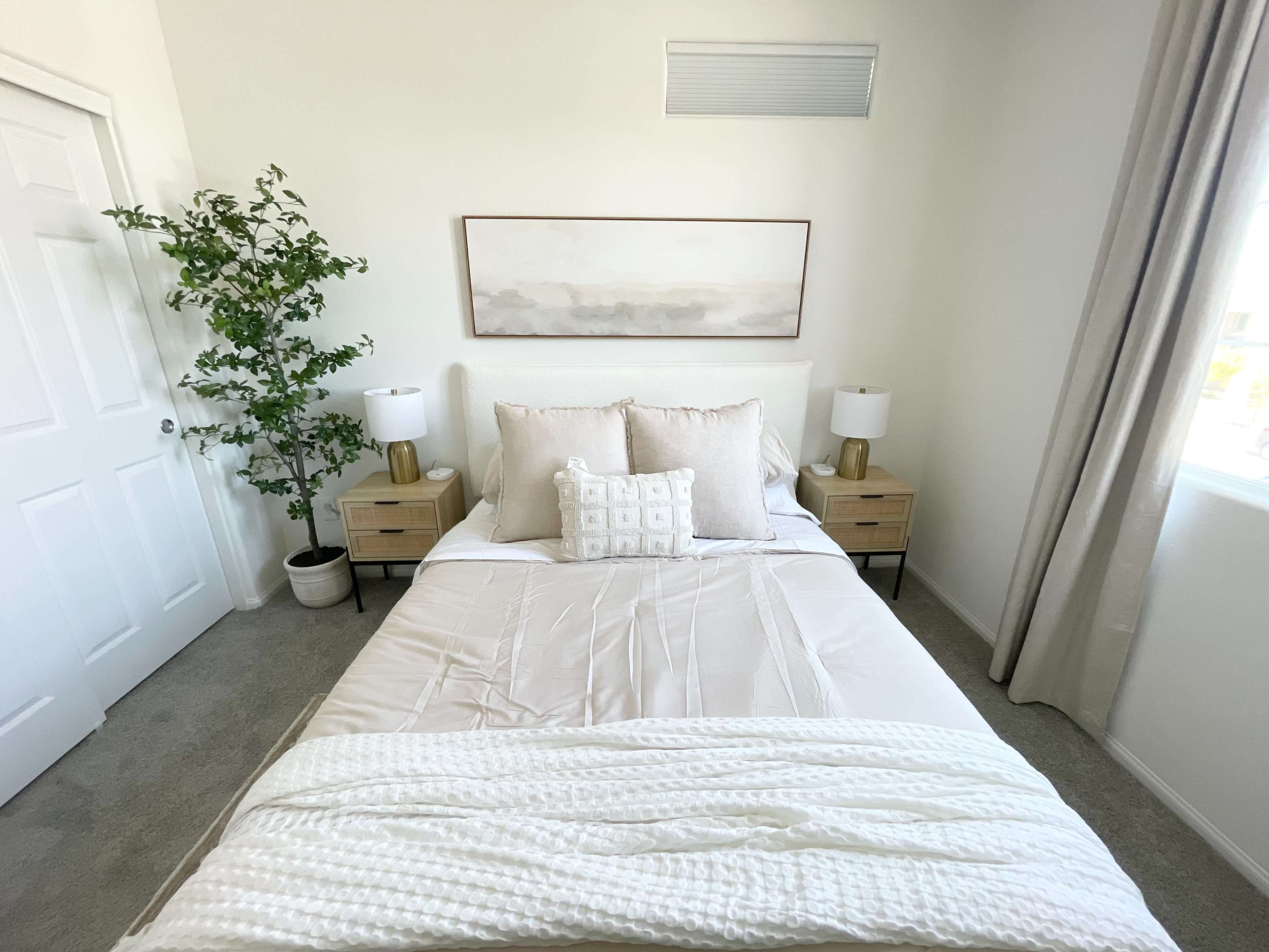 A neatly arranged bedroom features a bed with light bedding, two nightstands with lamps, and a decorative plant beside the window.