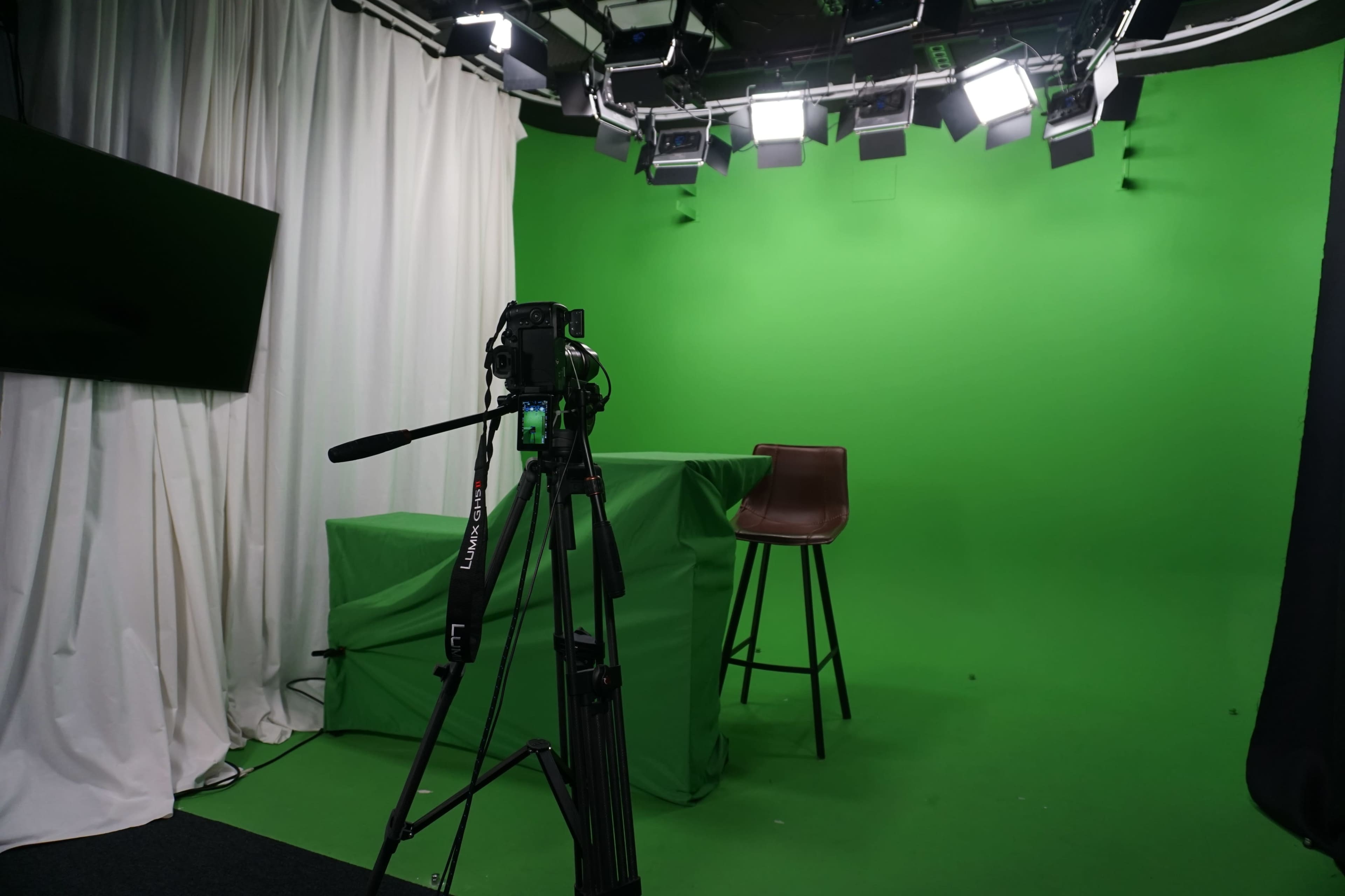 The image shows a green screen studio setup with a camera on a tripod facing an empty stool and a draped table in front of a green backdrop.