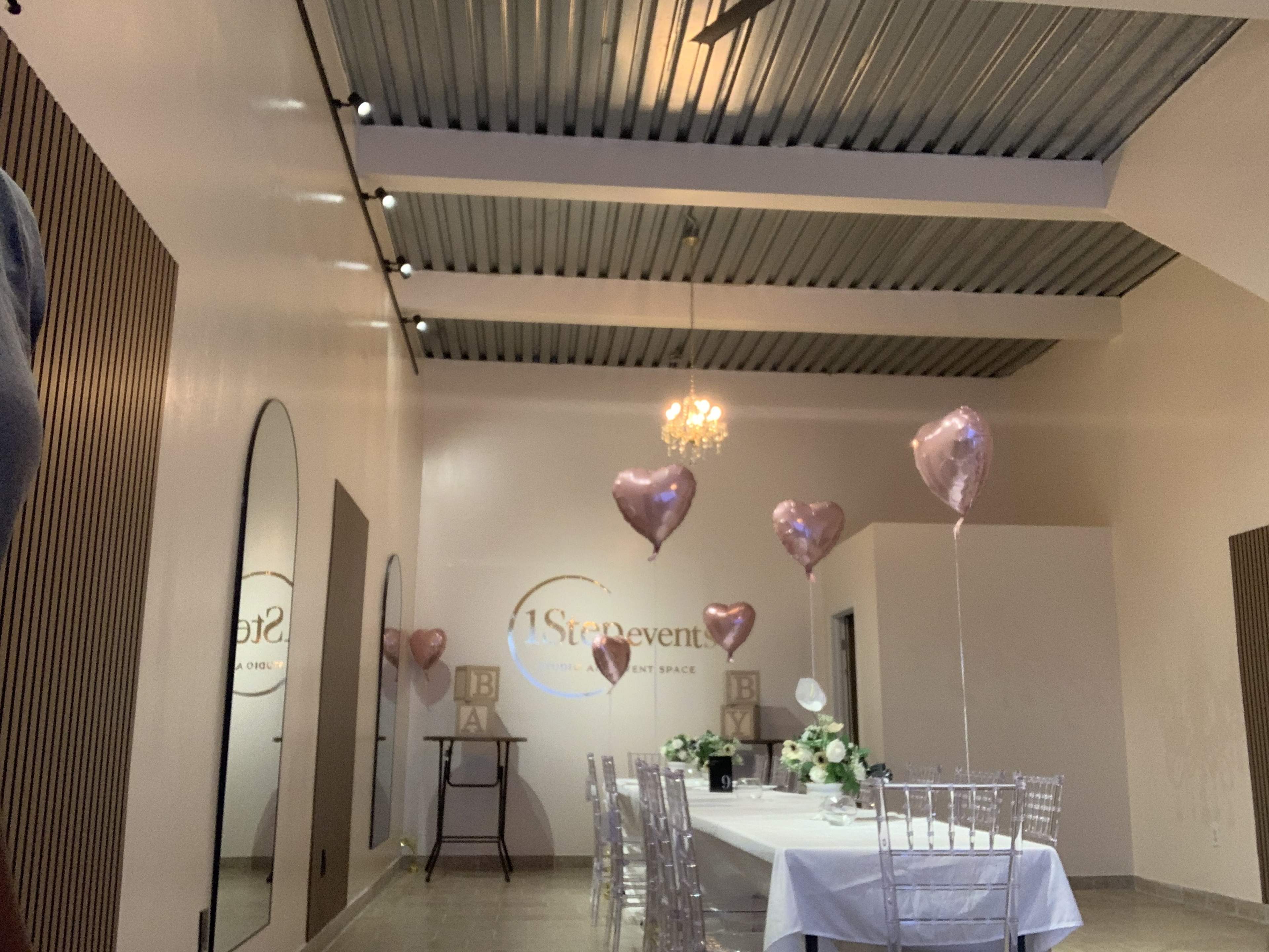 The image shows a well-decorated event space featuring a long table set for a gathering, with heart-shaped balloons and a chandelier above.