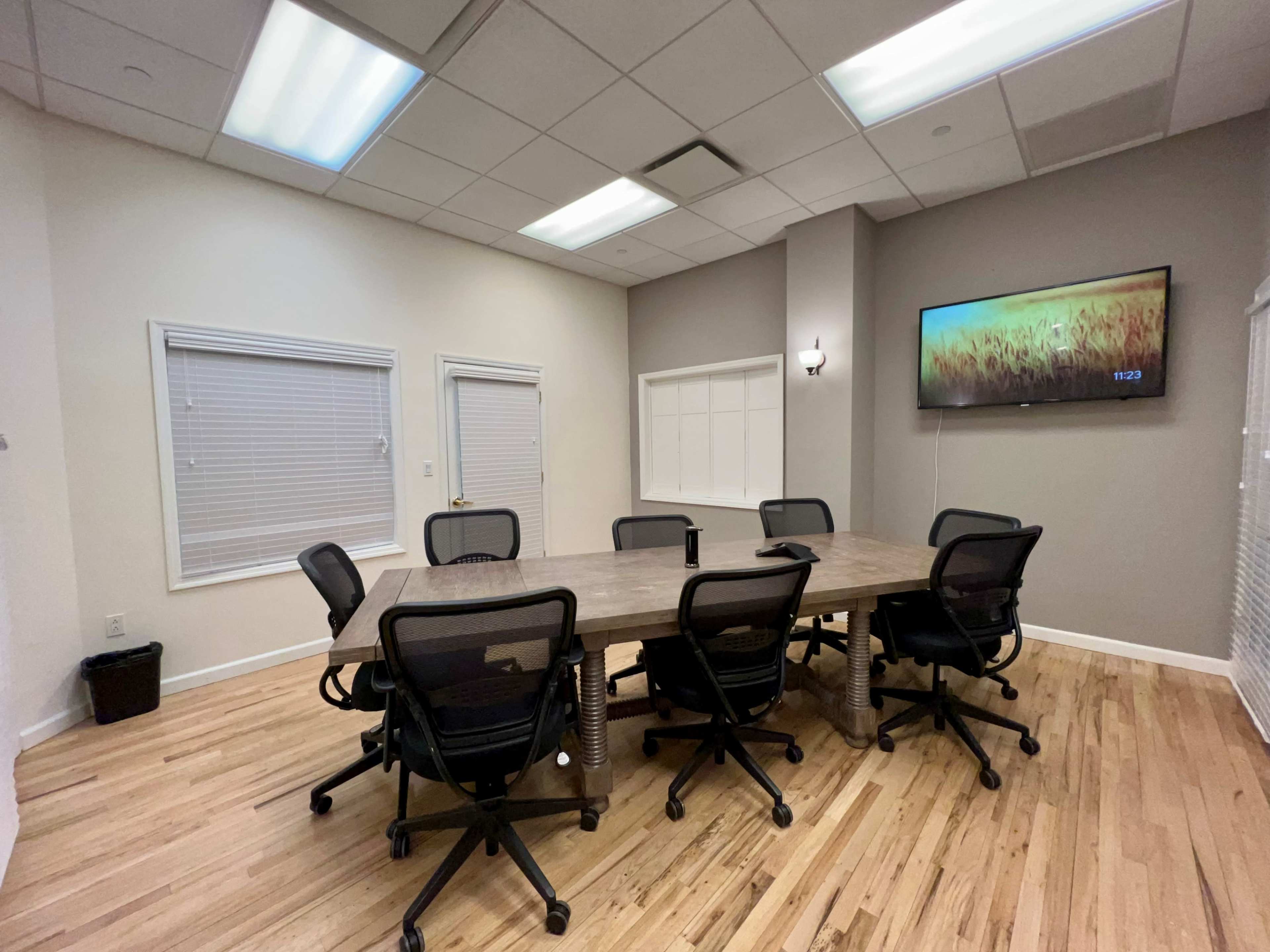 A conference room features a large wooden table surrounded by eight rolling chairs, with a TV mounted on the wall and windows with blinds.