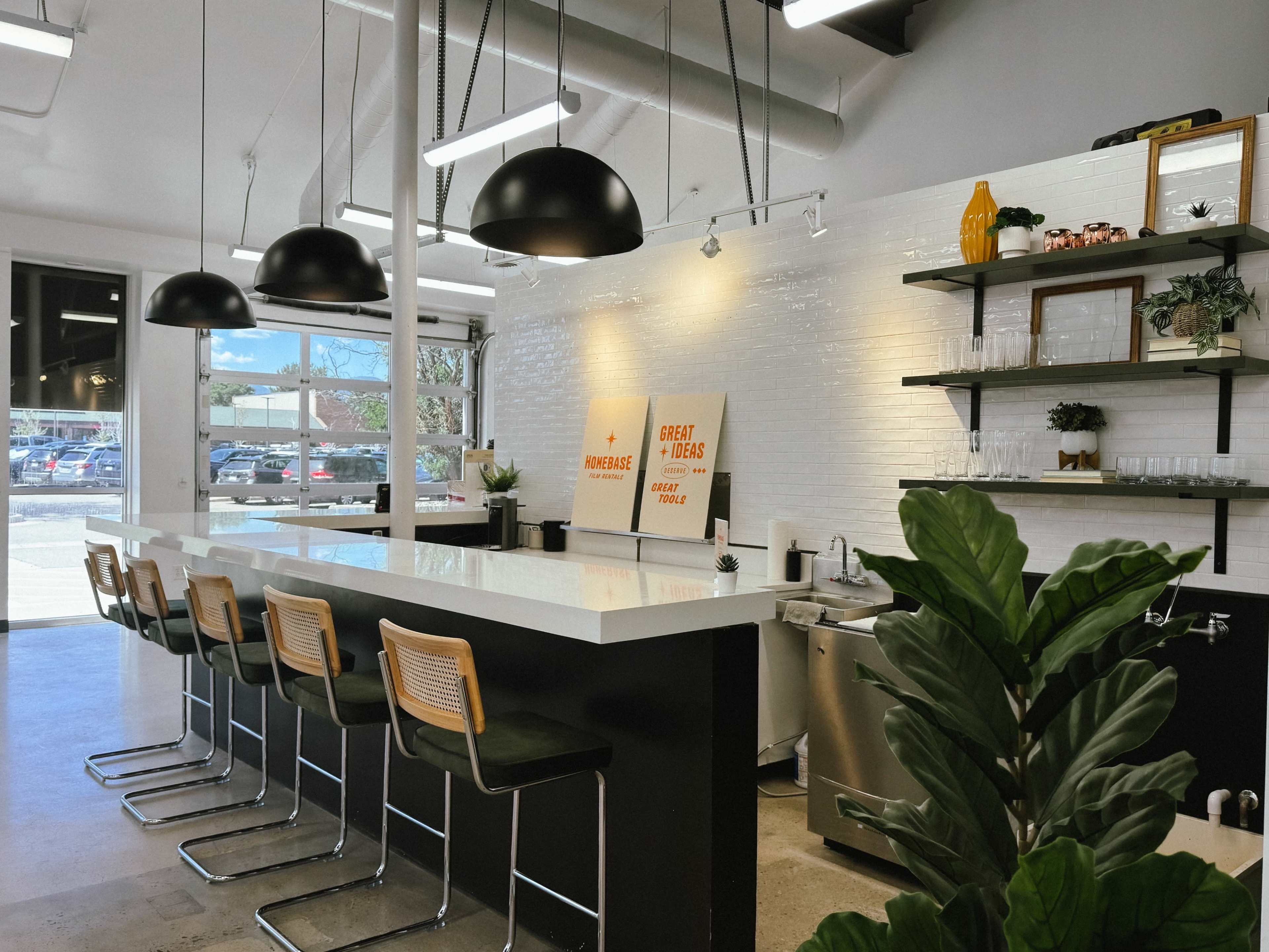 The image shows a modern café interior with a long white counter bar lined with black and wicker stools, pendant lights above, and shelves displaying plants and décor.