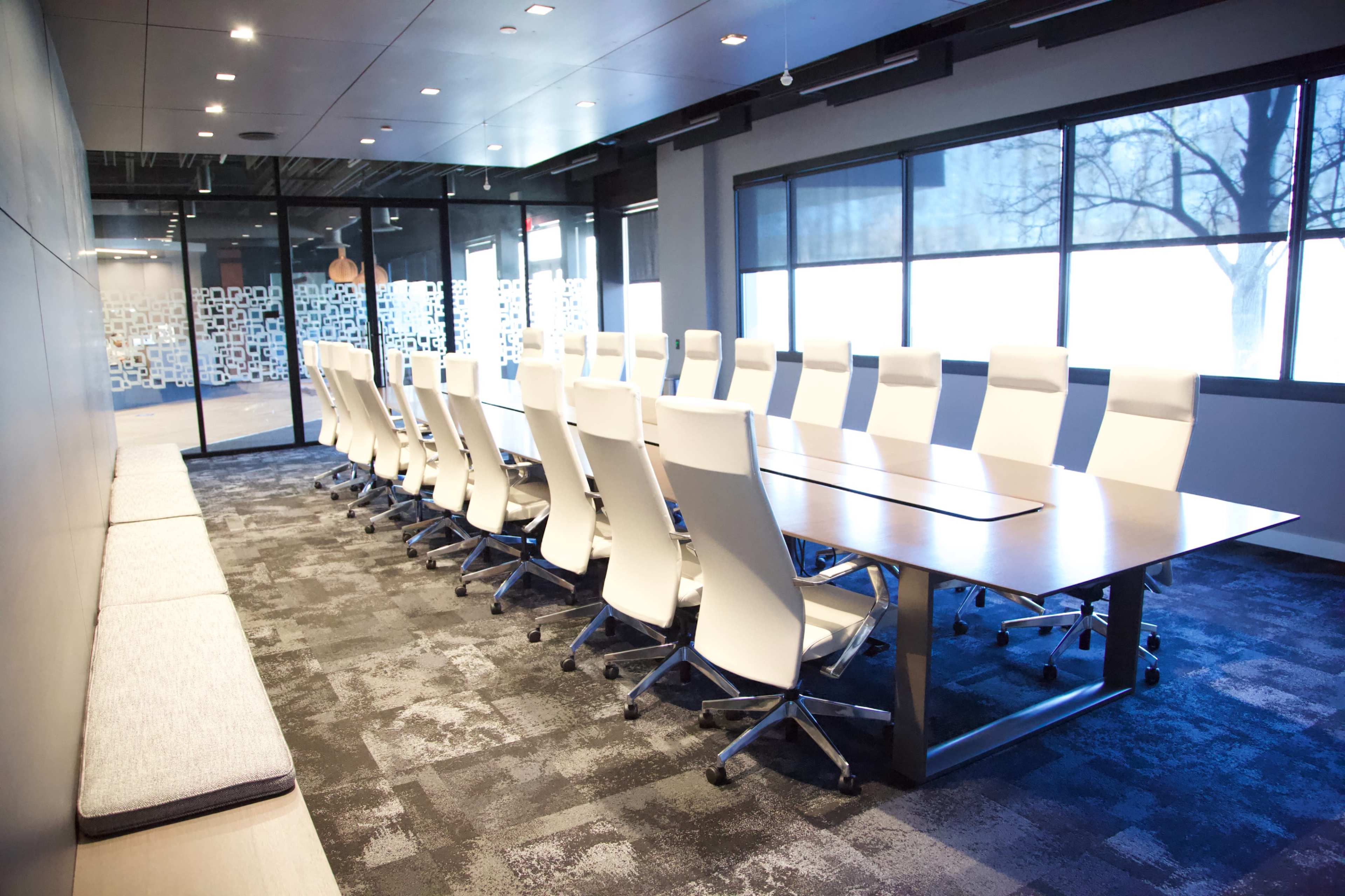 The image shows a modern conference room featuring a long table surrounded by white chairs, with large windows providing natural light.