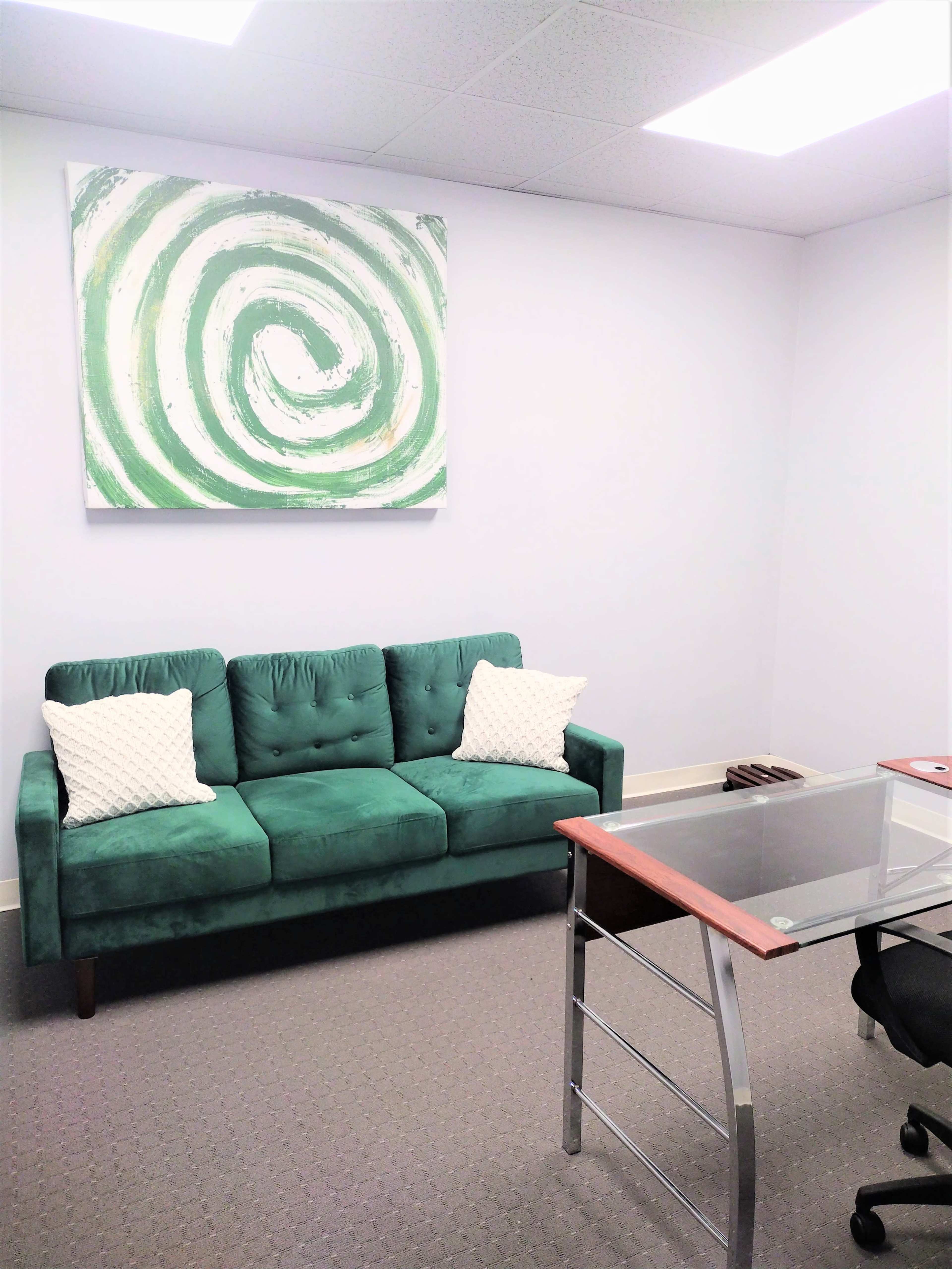 The image shows a small office space with a green couch adorned with two white pillows, a glass desk, and a large abstract swirl painting on the wall.