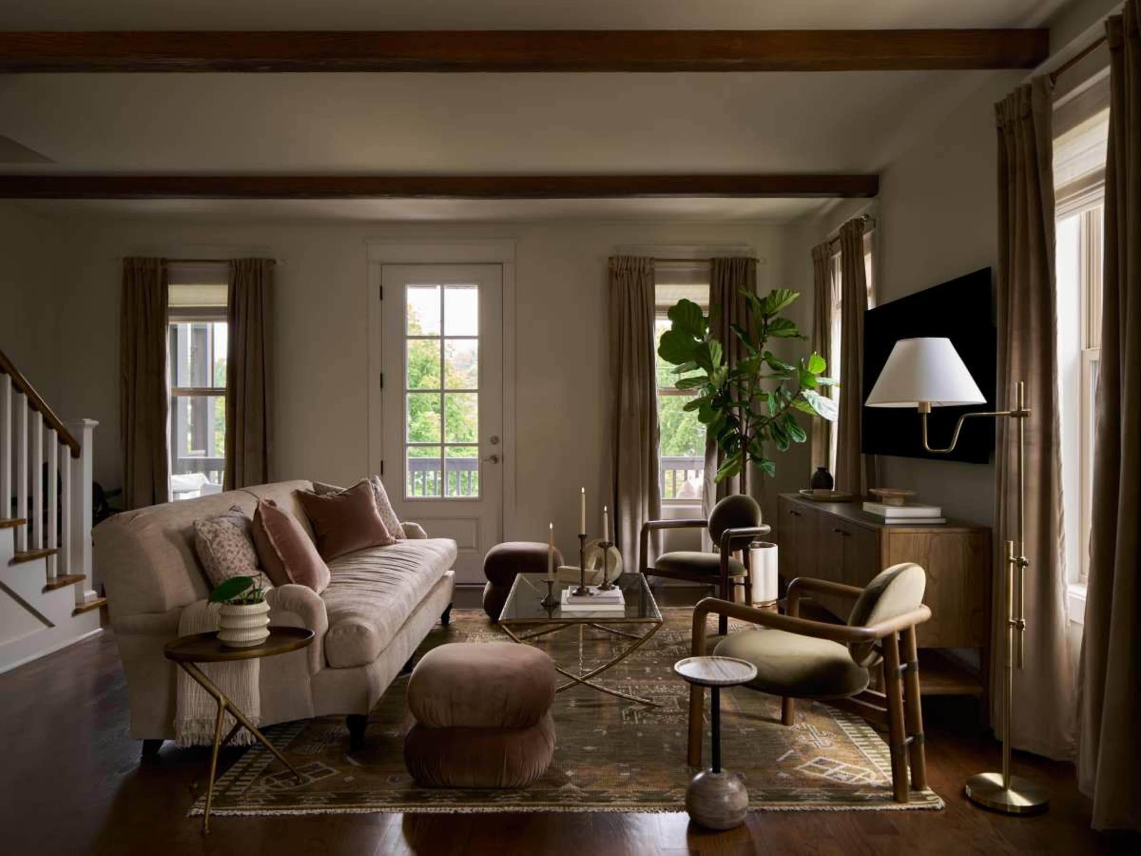 A cozy living room features a beige sofa, a glass coffee table, and large windows with sheer curtains, complemented by plants and decorative accessories.