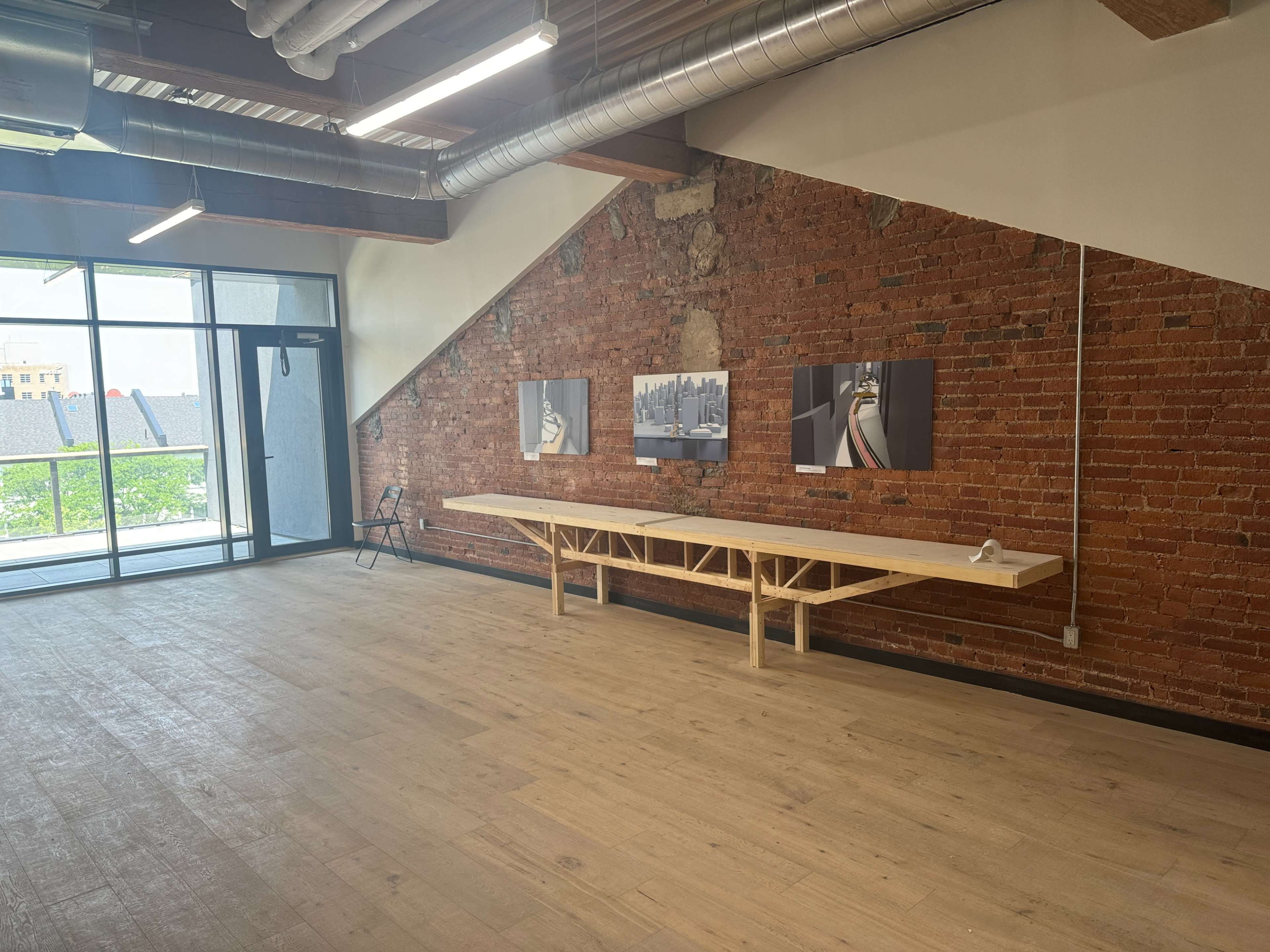 A spacious room with a wooden table along a brick wall, featuring two framed pictures, and large windows allowing natural light to enter.