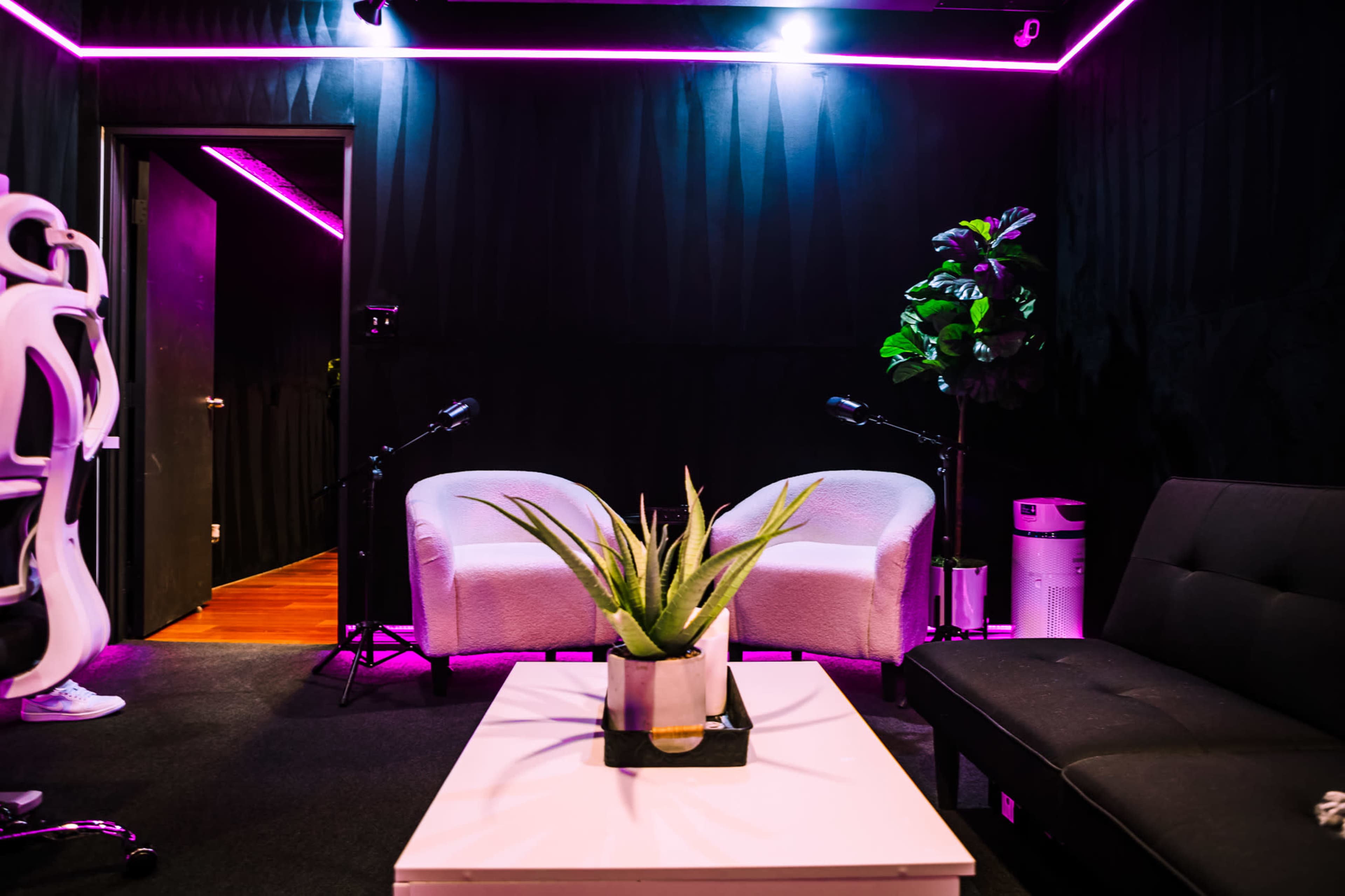 A cozy room features two white chairs, a black futon, a small plant on a coffee table, and a microphone setup under pink neon lighting.