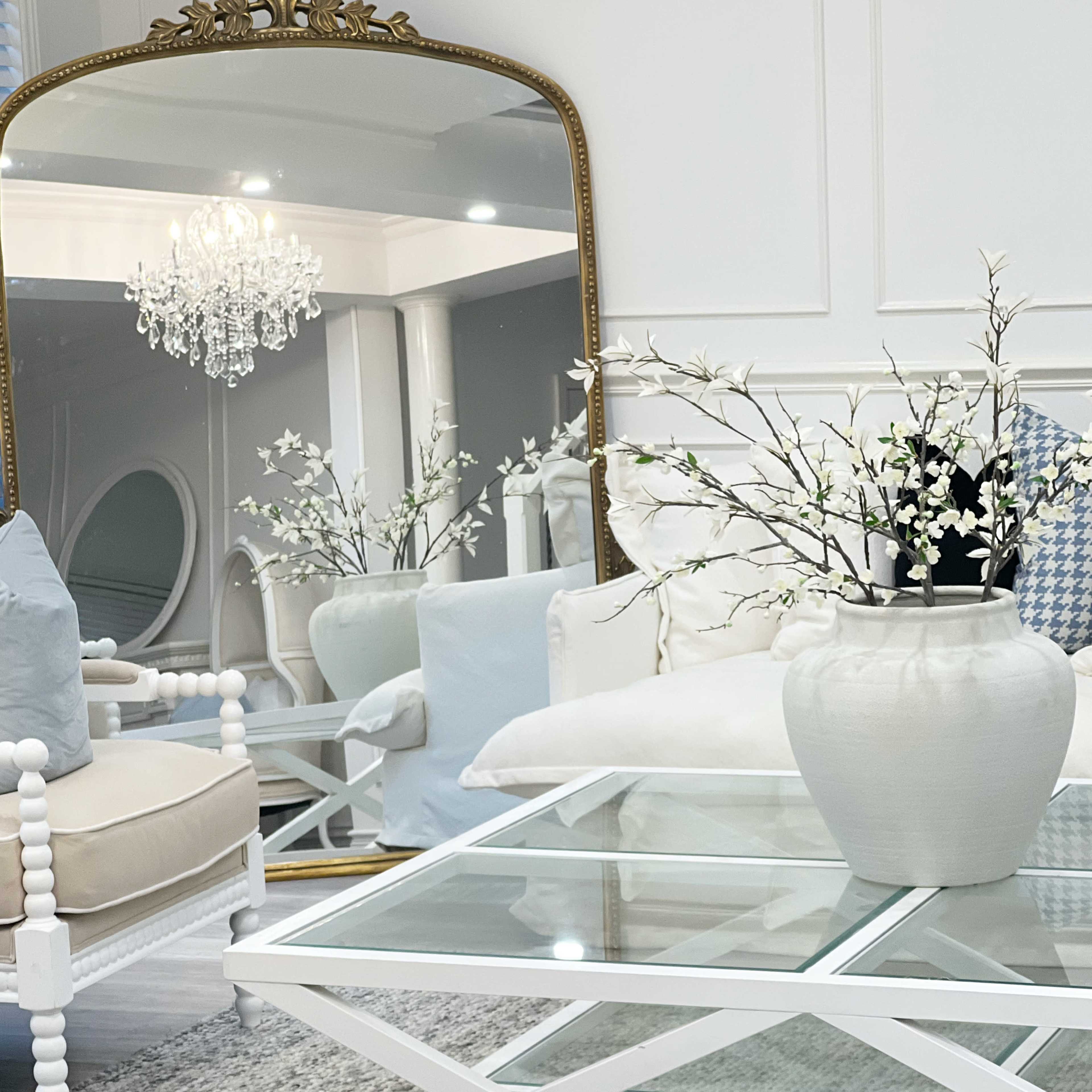 A living room features a large mirror reflecting a chandelier, with a white vase of flowers atop a glass coffee table and stylish seating.