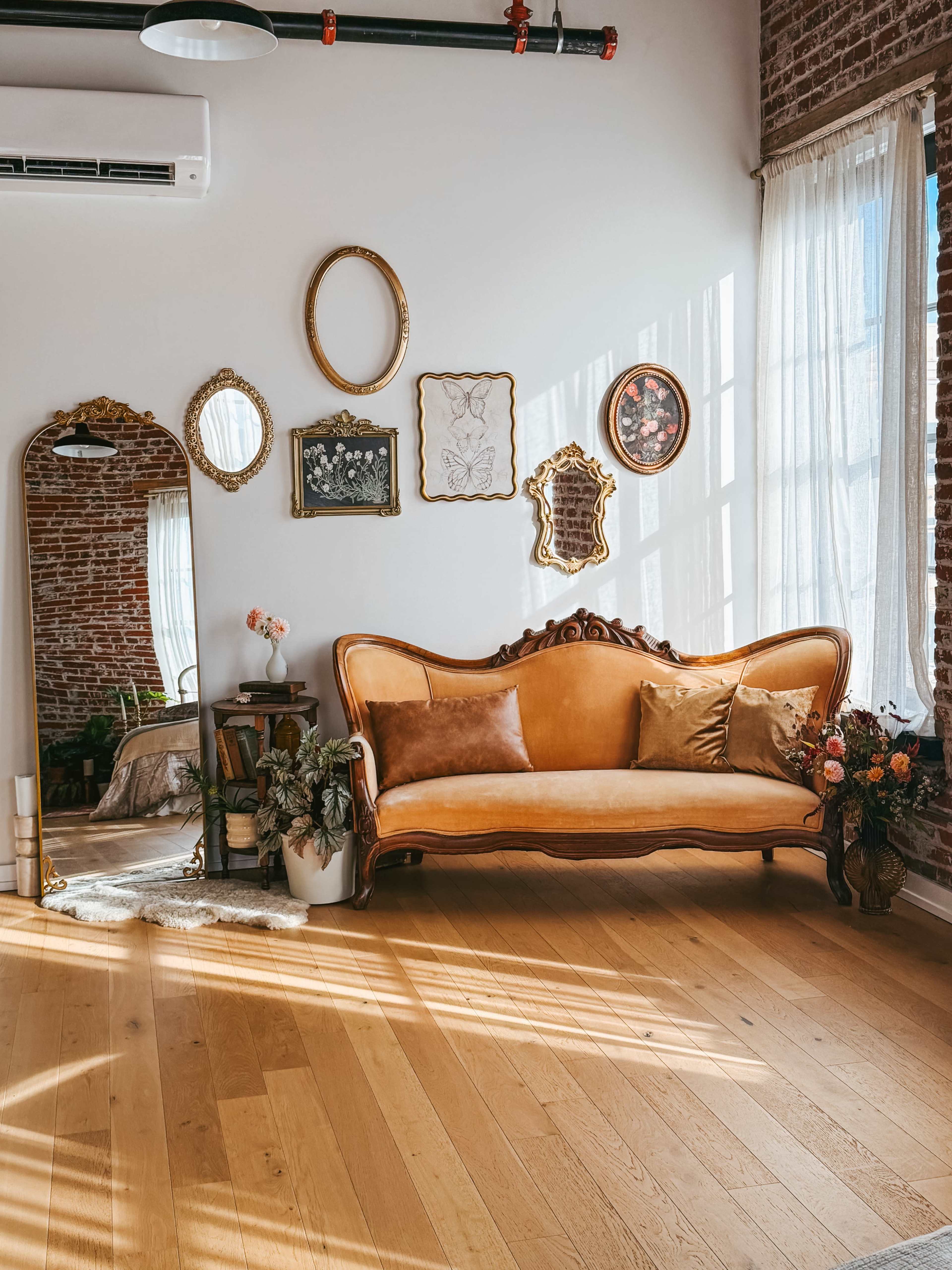 A vintage sofa sits against a brick wall, surrounded by decorative mirrors and framed artwork, with sunlight streaming through large windows.