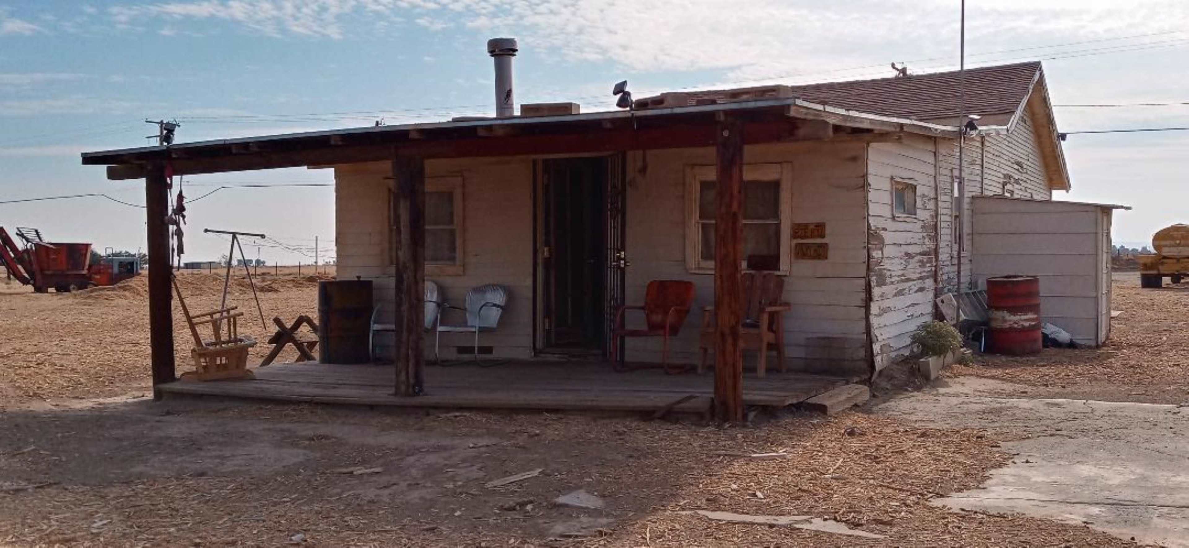 An old, weathered house with a wooden porch and a few chairs, set in a rural landscape with dry ground.
