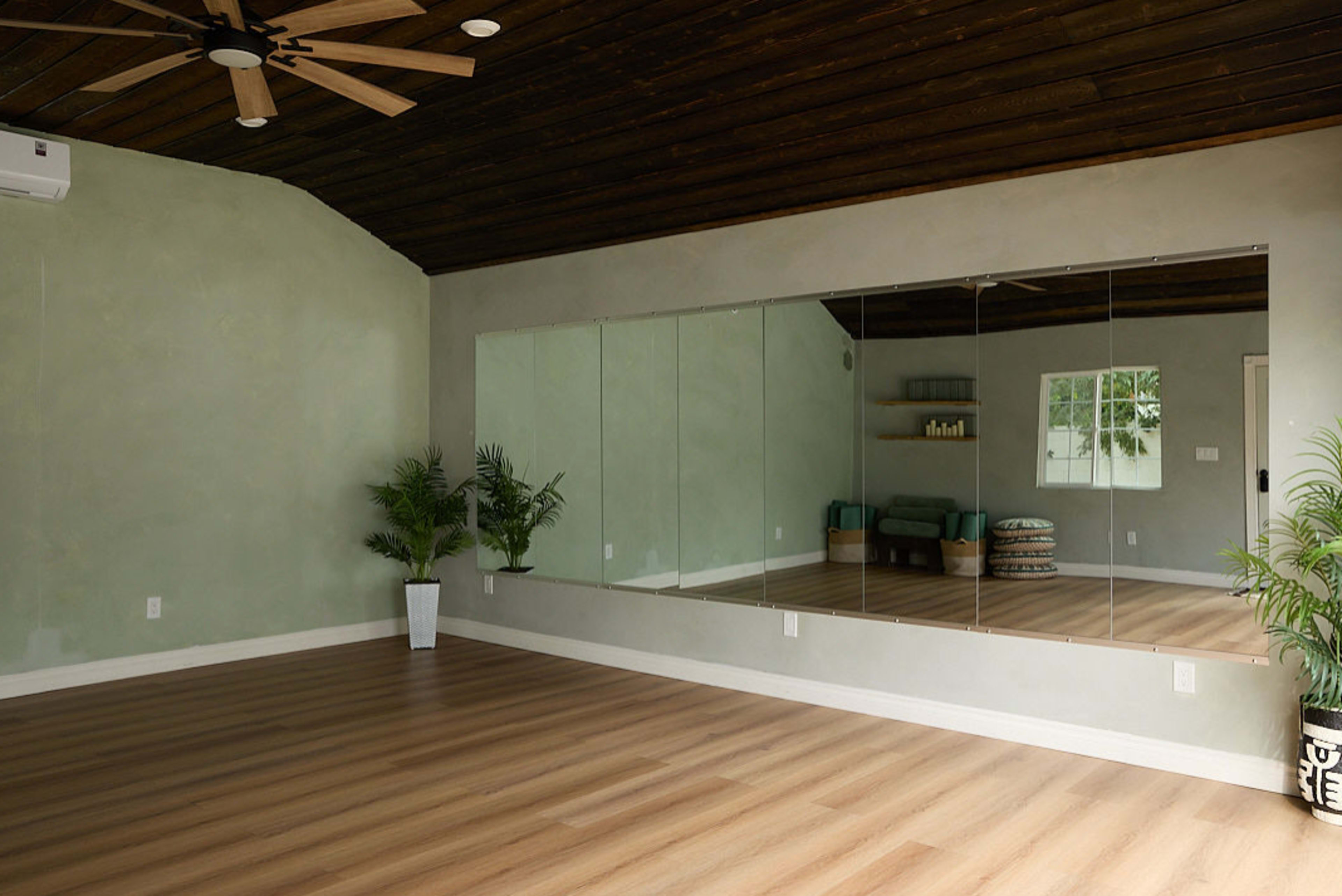 The image shows a spacious room with a wooden ceiling, a large mirror on one wall, and simple furnishings including a couch and decorative plants.