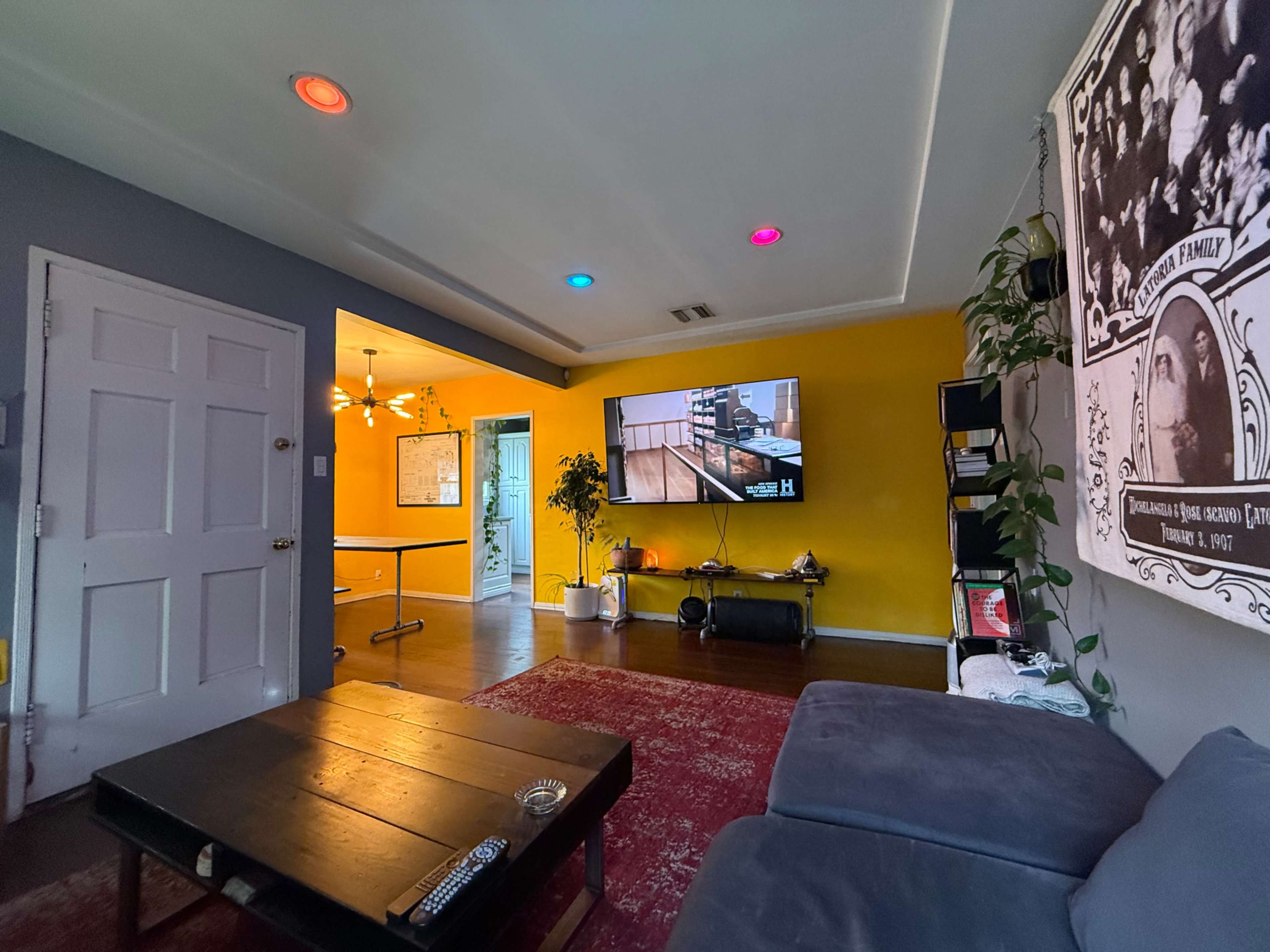 A living room features a gray wall with a large television mounted above a black entertainment unit, adjacent to a bright yellow wall and a doorway leading to another space.