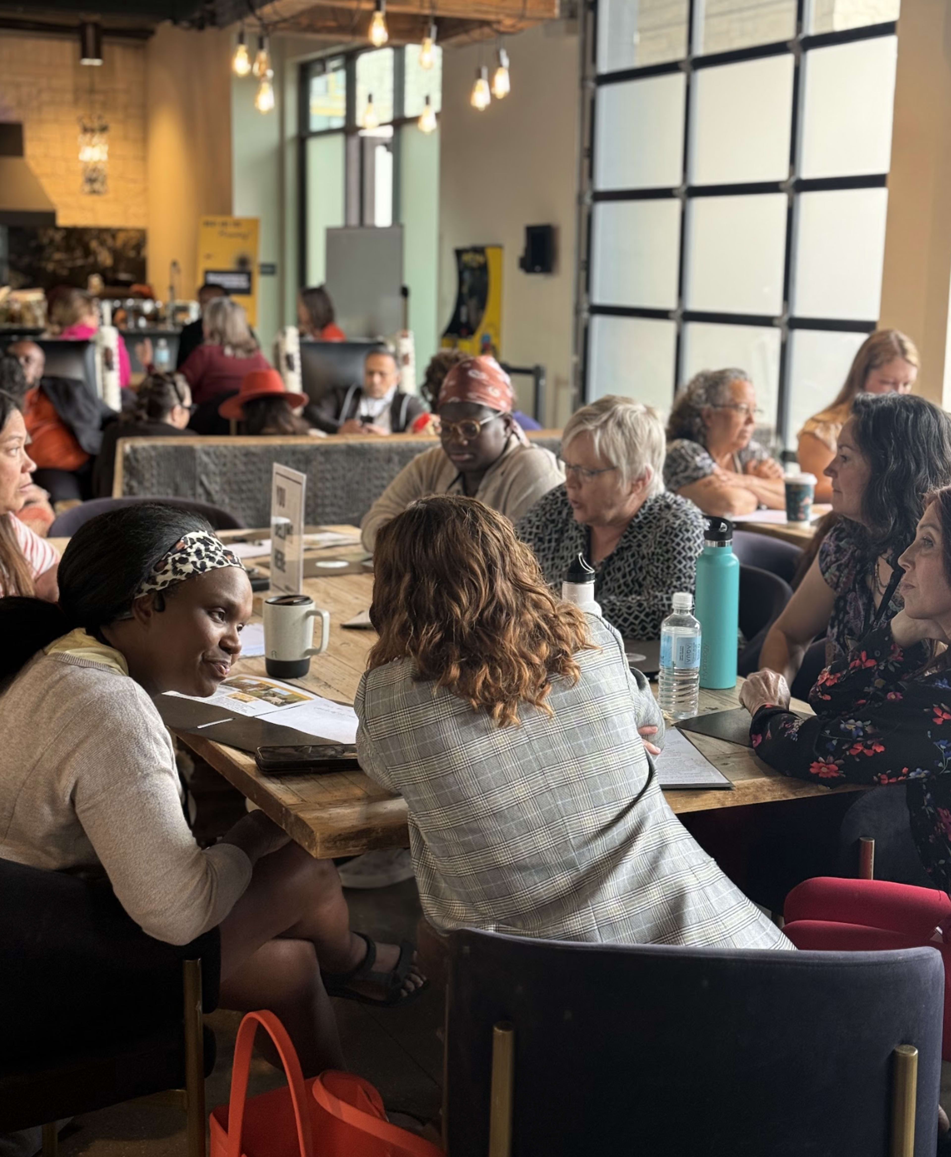 A group of people sits around tables in a café, engaged in discussion and collaboration.