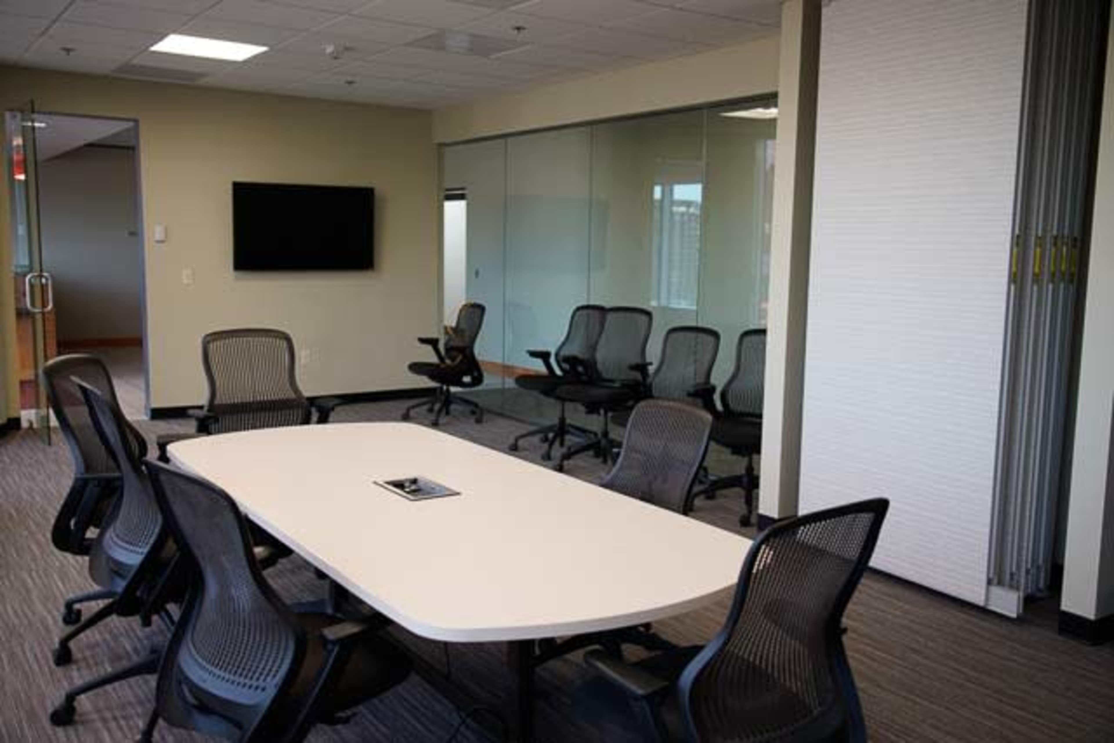 A conference room features a large white table surrounded by black office chairs, with a television mounted on the wall and glass partitions.