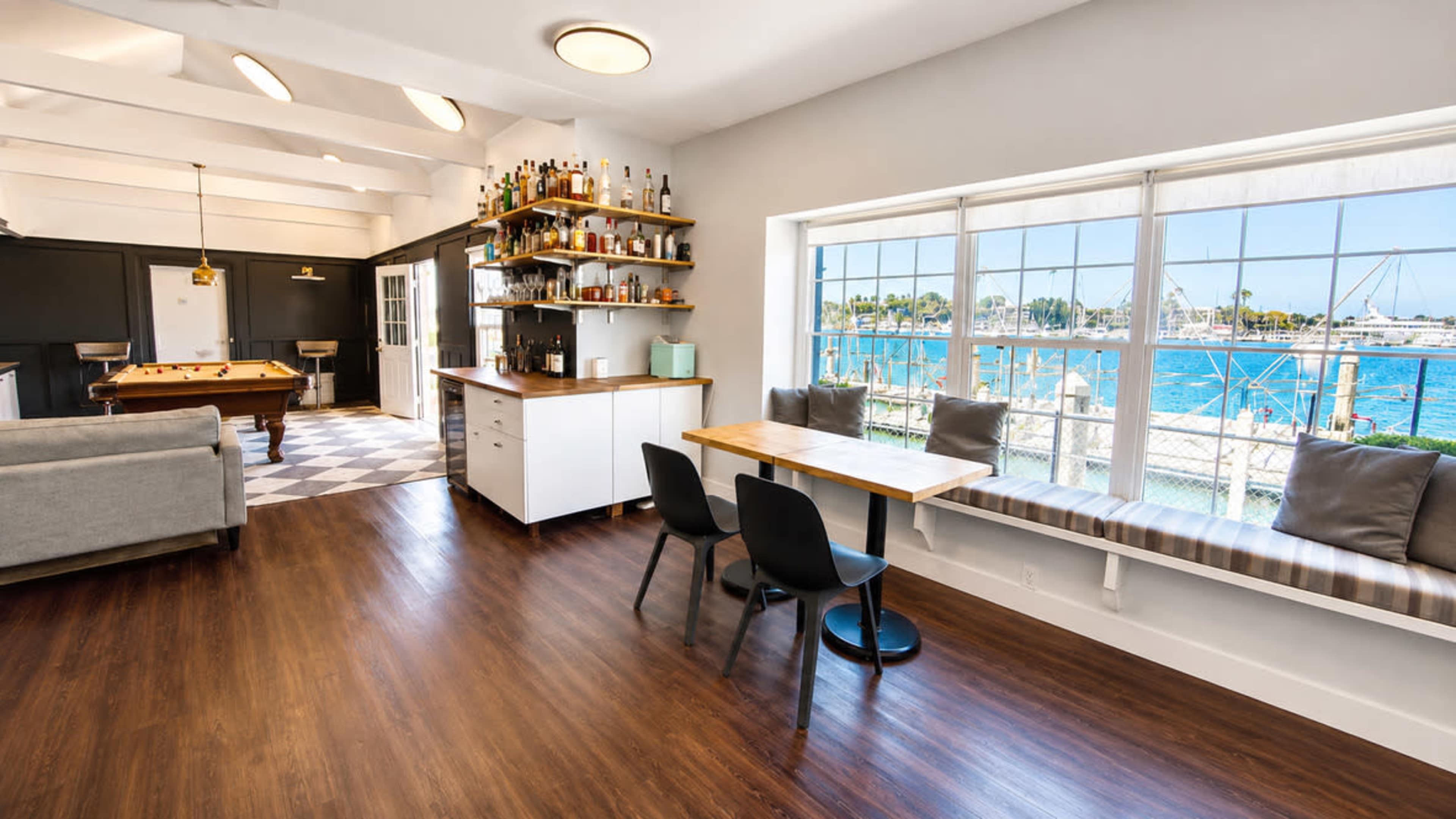 The image shows a bright, modern interior with a seating area, a pool table, and expansive windows overlooking a harbor.