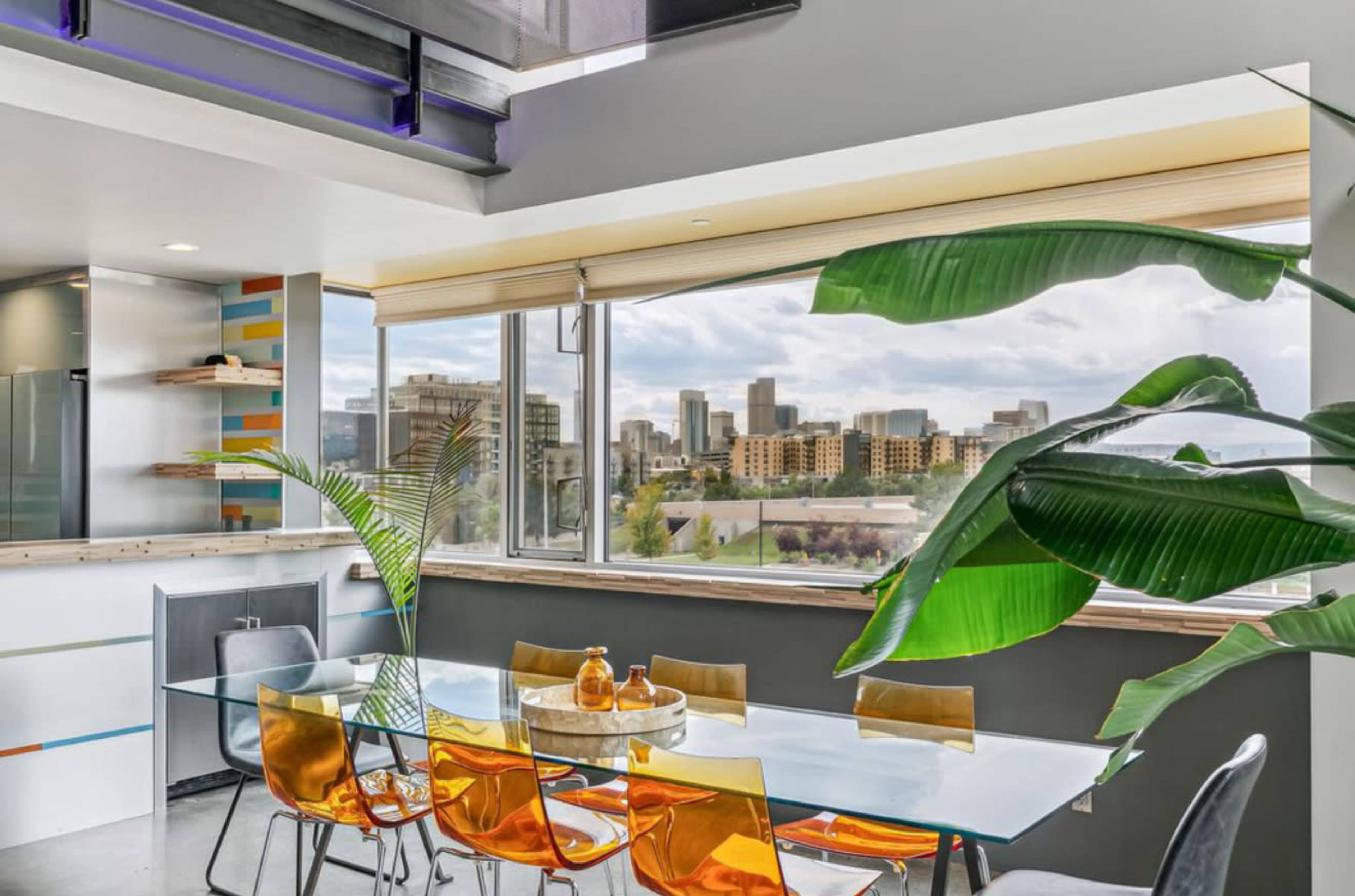 A modern dining area features a glass table with colorful chairs, surrounded by a large window that provides a view of a city skyline.