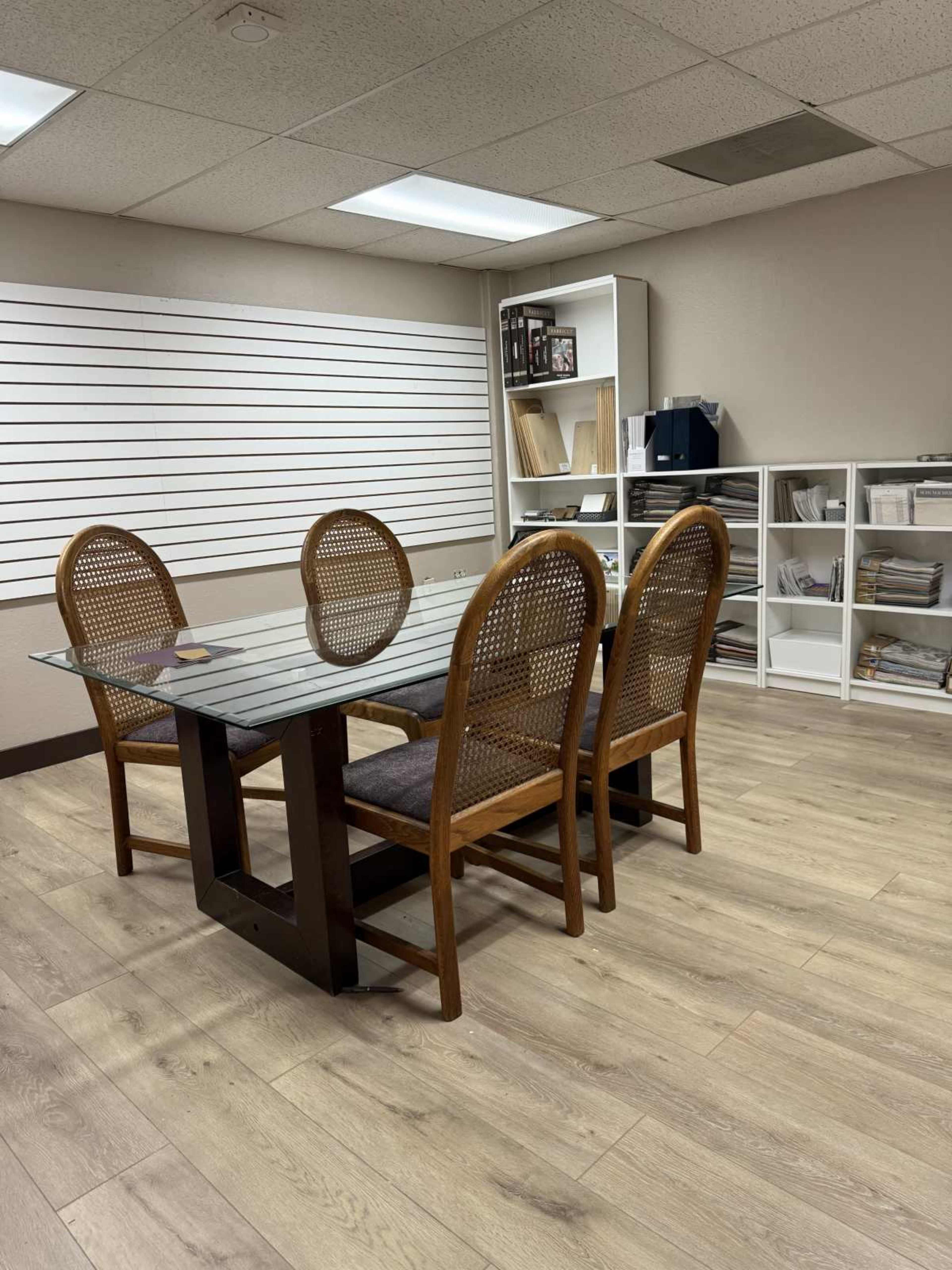 The image shows a small meeting room with a glass-top table and four wooden chairs, accompanied by shelving units filled with documents and materials.