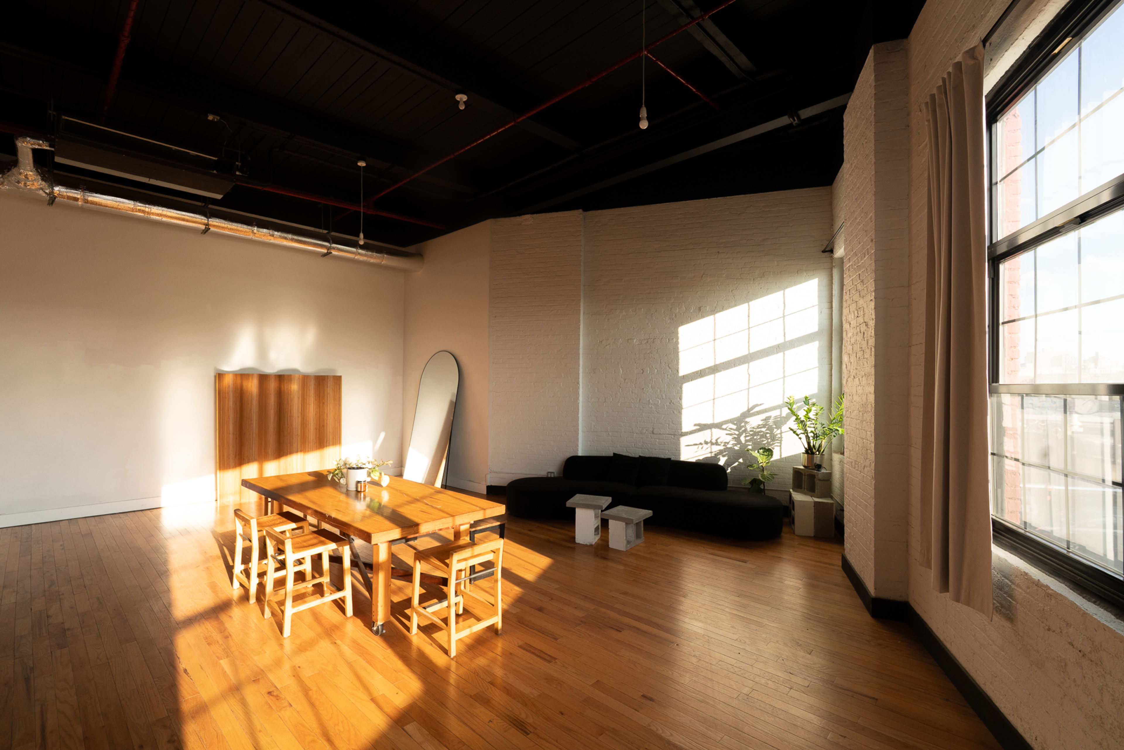 The image shows a spacious, sunlit room with a wooden dining table and chairs, a black sofa, a large mirror, and a plant near a window.