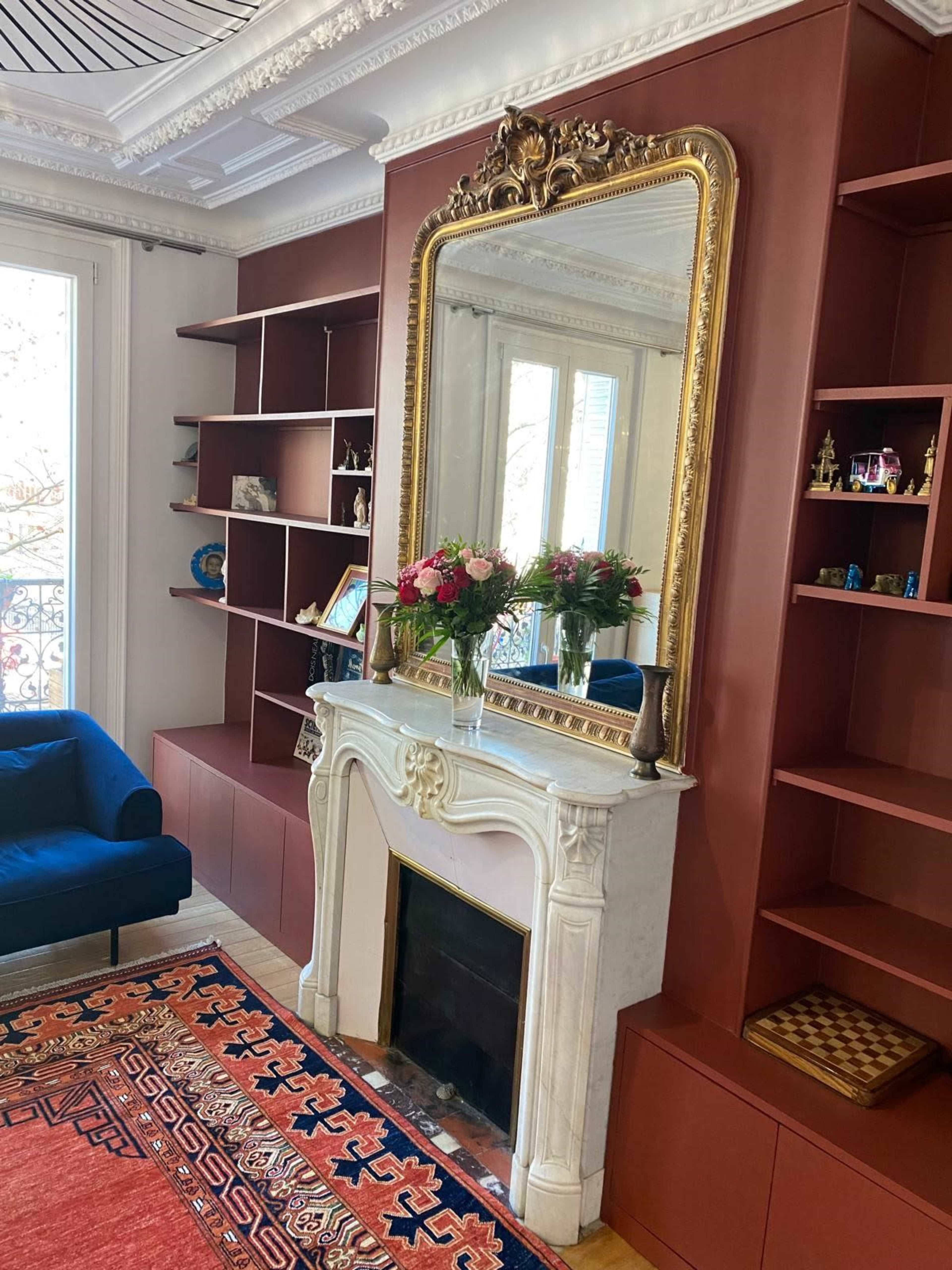 The image shows a living room with a white marble fireplace, a large ornate mirror above it, a blue sofa, and red shelving units against the walls, adorned with various decorative items and a bouquet of flowers on the mantel.