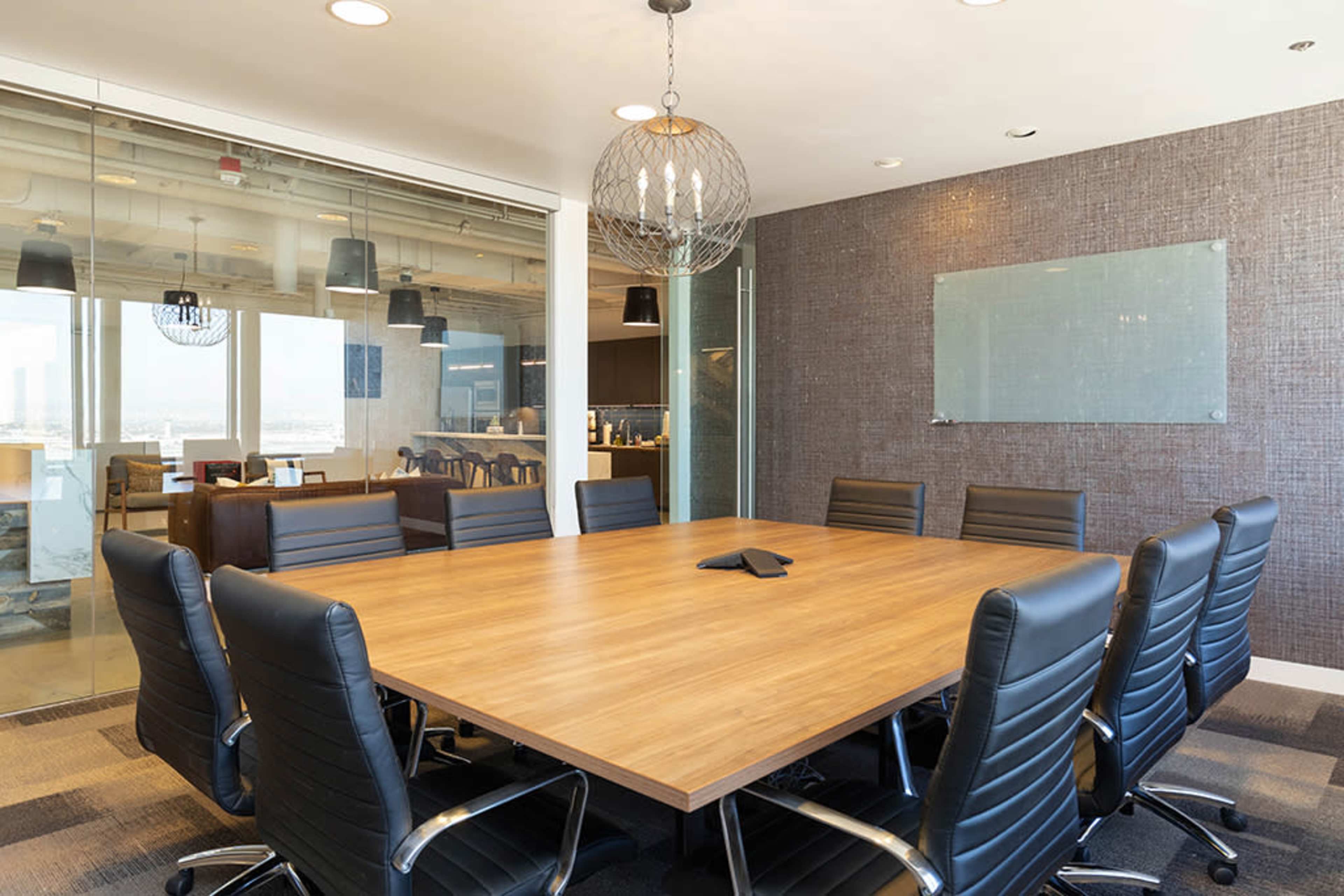 A modern conference room features a large wooden table surrounded by black chairs and a glass wall that separates it from another office area.